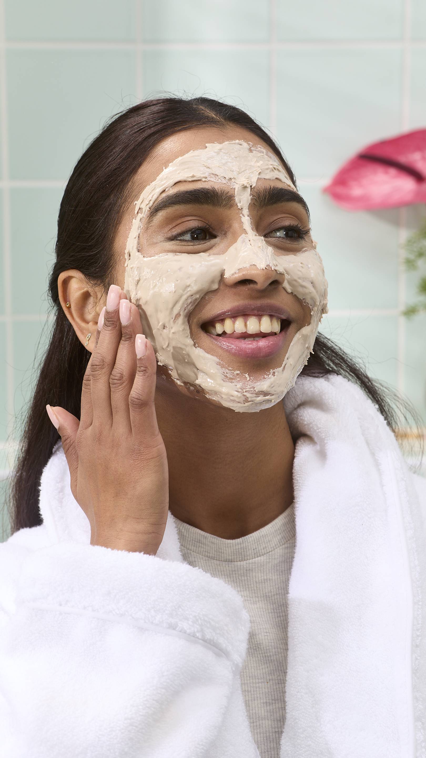 A model with dark hair tied back smiles as they apply a thick, beige-coloured face mask over their face, leaving only their eyes and lips clear. They wear a white fluffy dressing gown against a background of pale green tiles.