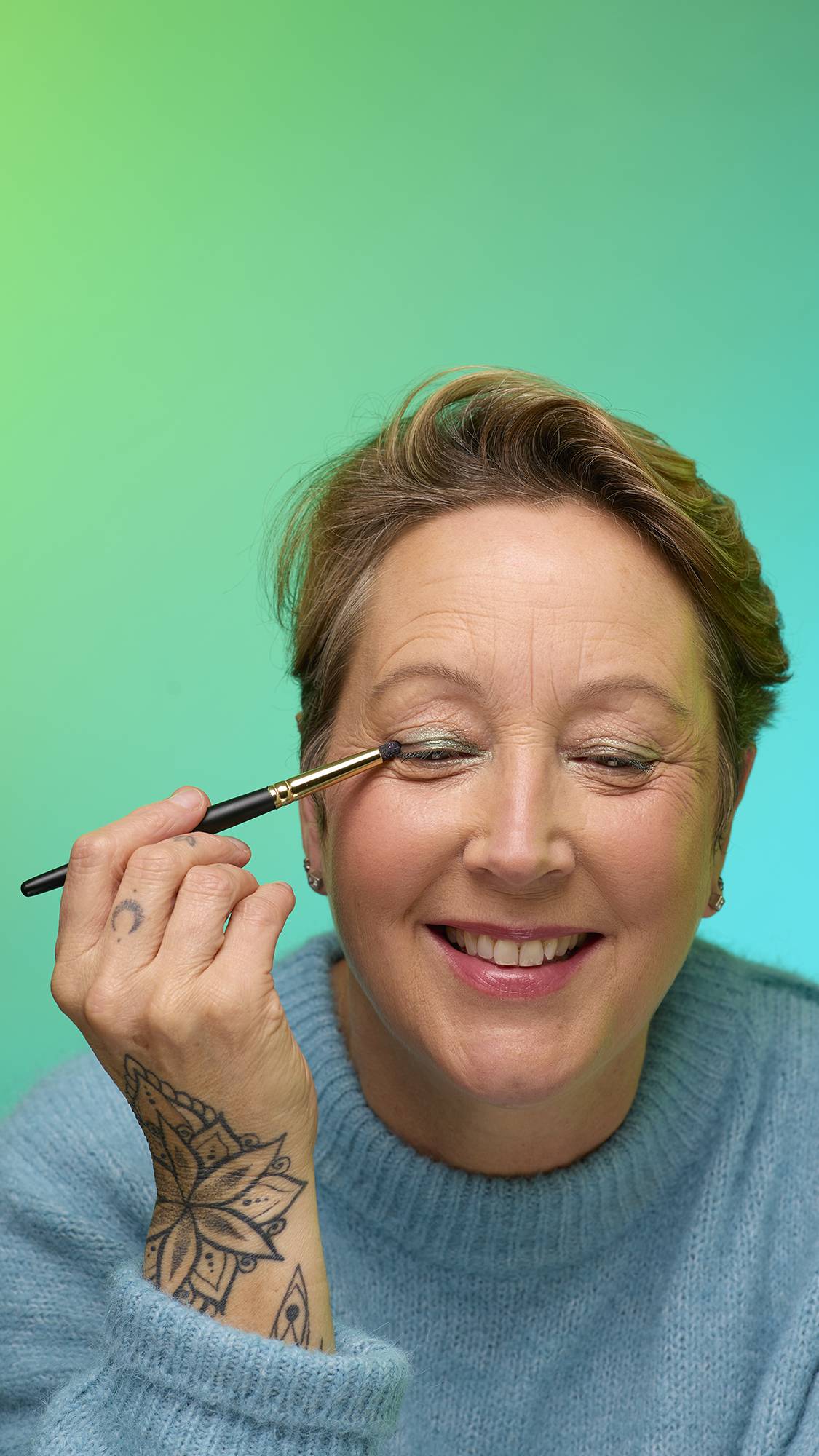 The image shows a close-up of the model's face on a pale blue-to-green gradient background. They are holding a thin make-up brush to apply a subtle, deep-sage-green, shimmering eyeshadow of the Meteor Glitter Crumble.
