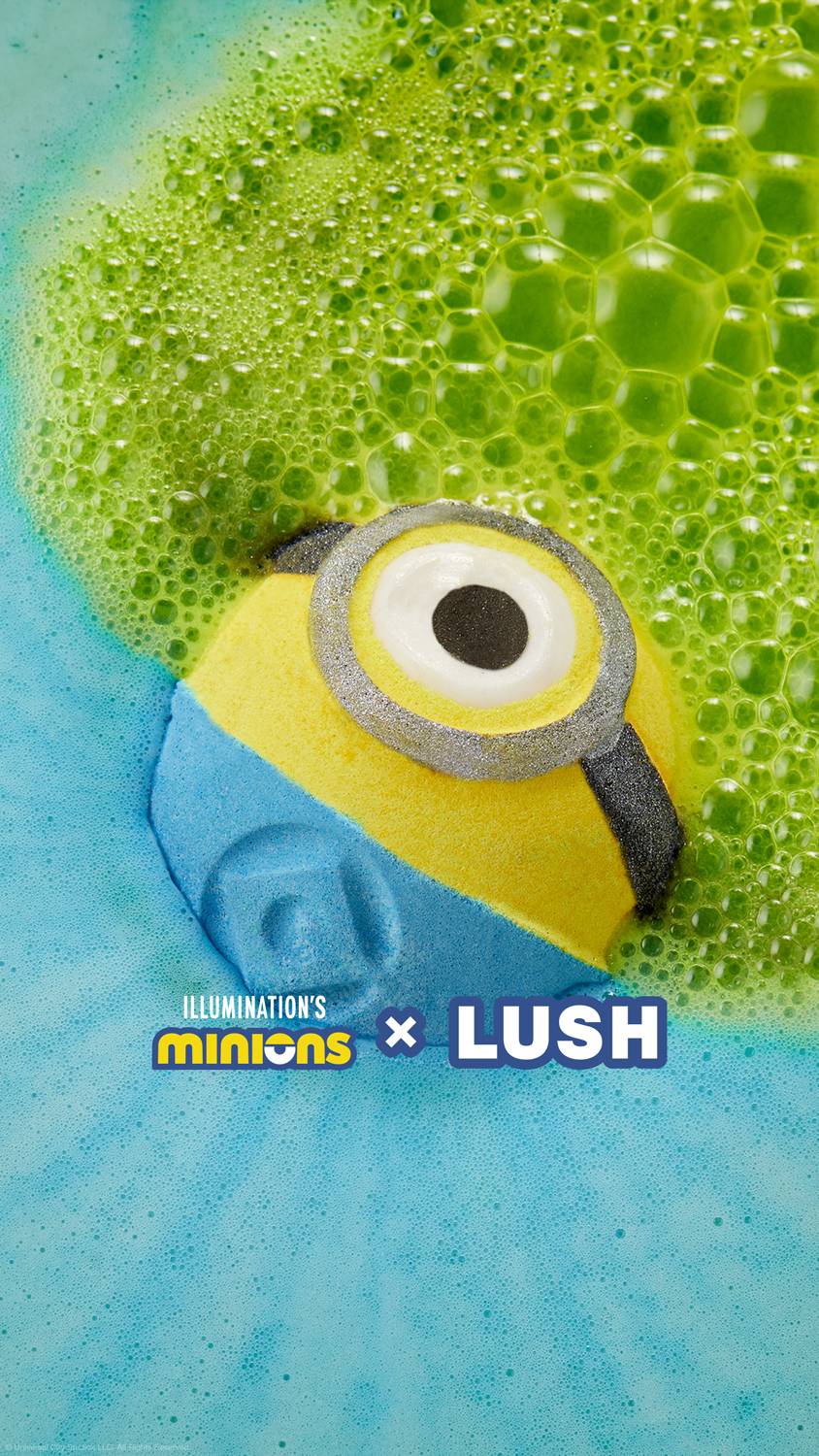 The Minions bath bomb is floating on the bath water as it starts to give off deep blue foam from the lower half and neon green foam from the top half. The Minions X LUSH logo is at the bottom.