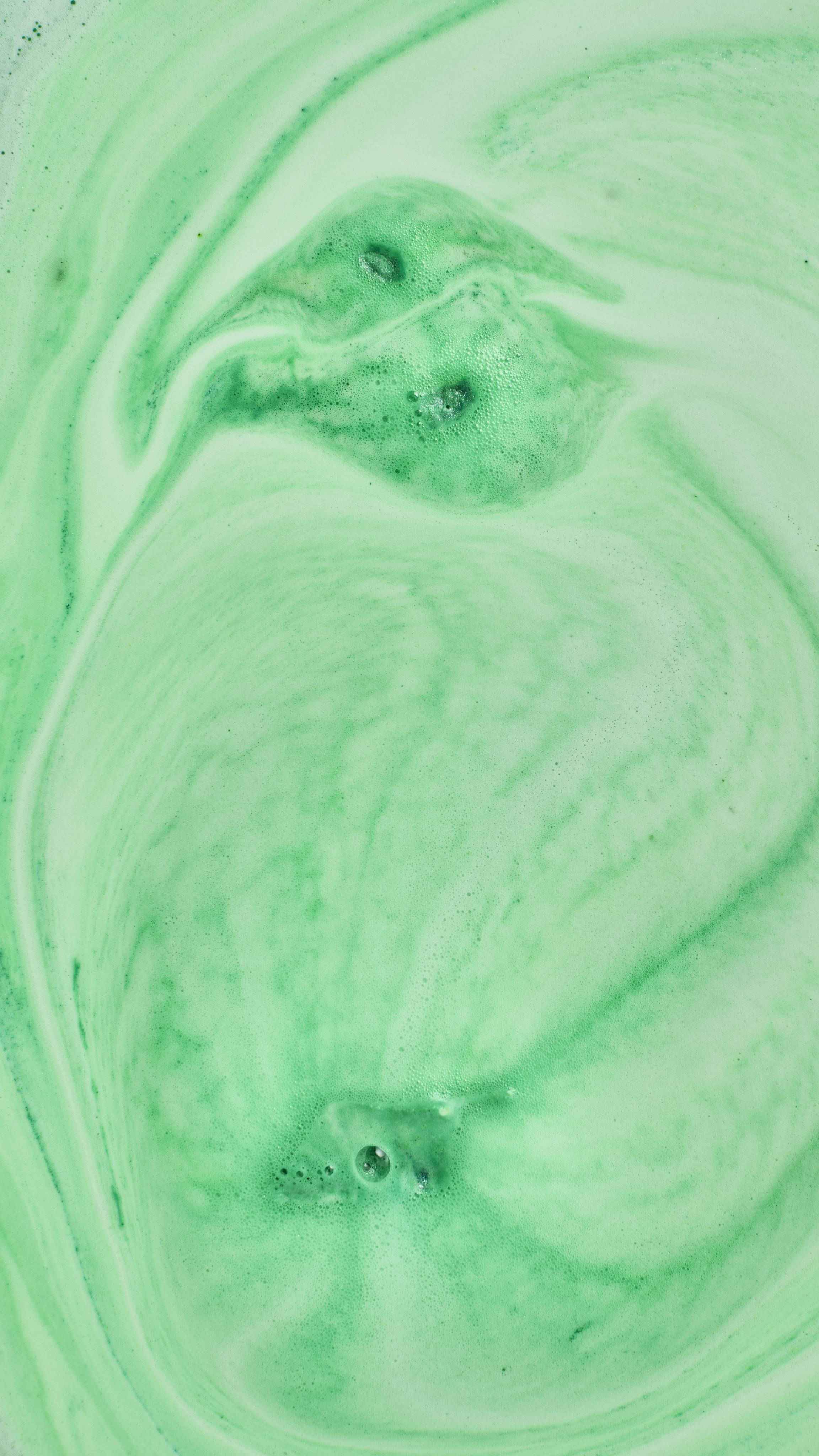 An abstract aerial shot of bath bomb foam swirling in water, creating marbled, wavy patterns of vibrant lime green and pale mint green. A few small clusters of soapy bubbles are scattered across the frothy surface.