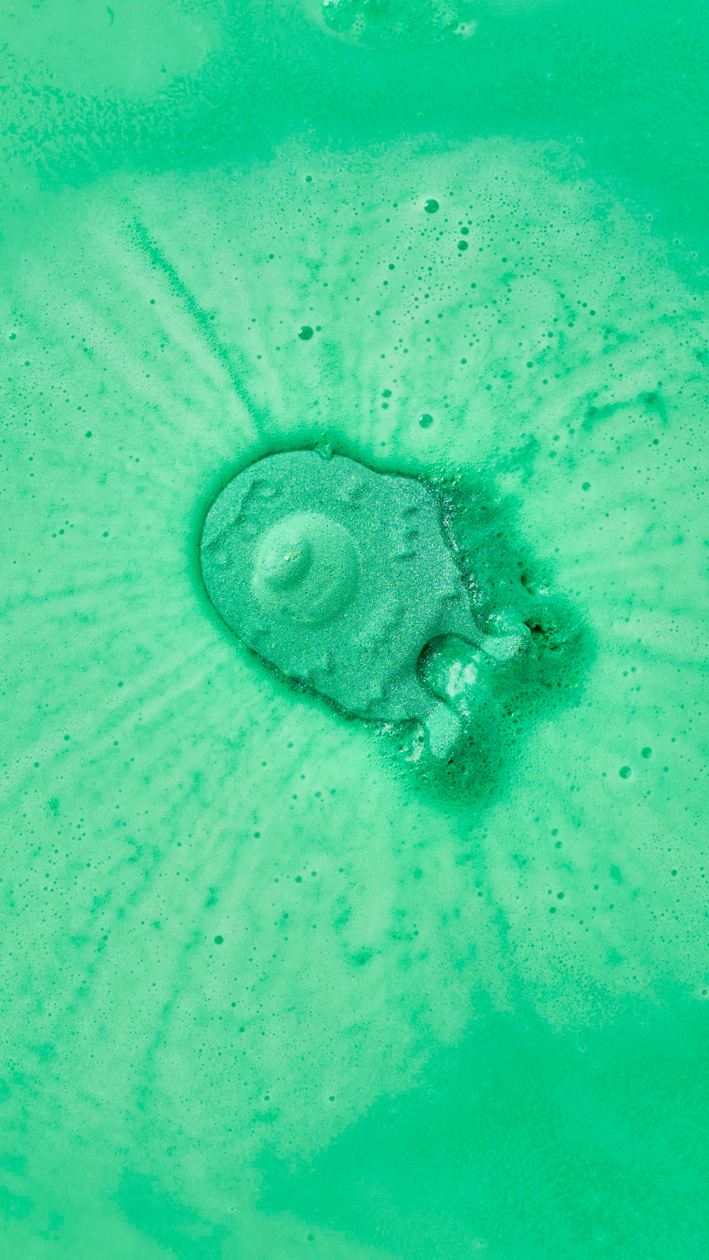 A mint-green, monster-shaped bath bomb dissolves at the centre of a bath, generating concentric ripples of lime-green and pale-green foam. The surrounding water is a vivid, opaque shade of green.
