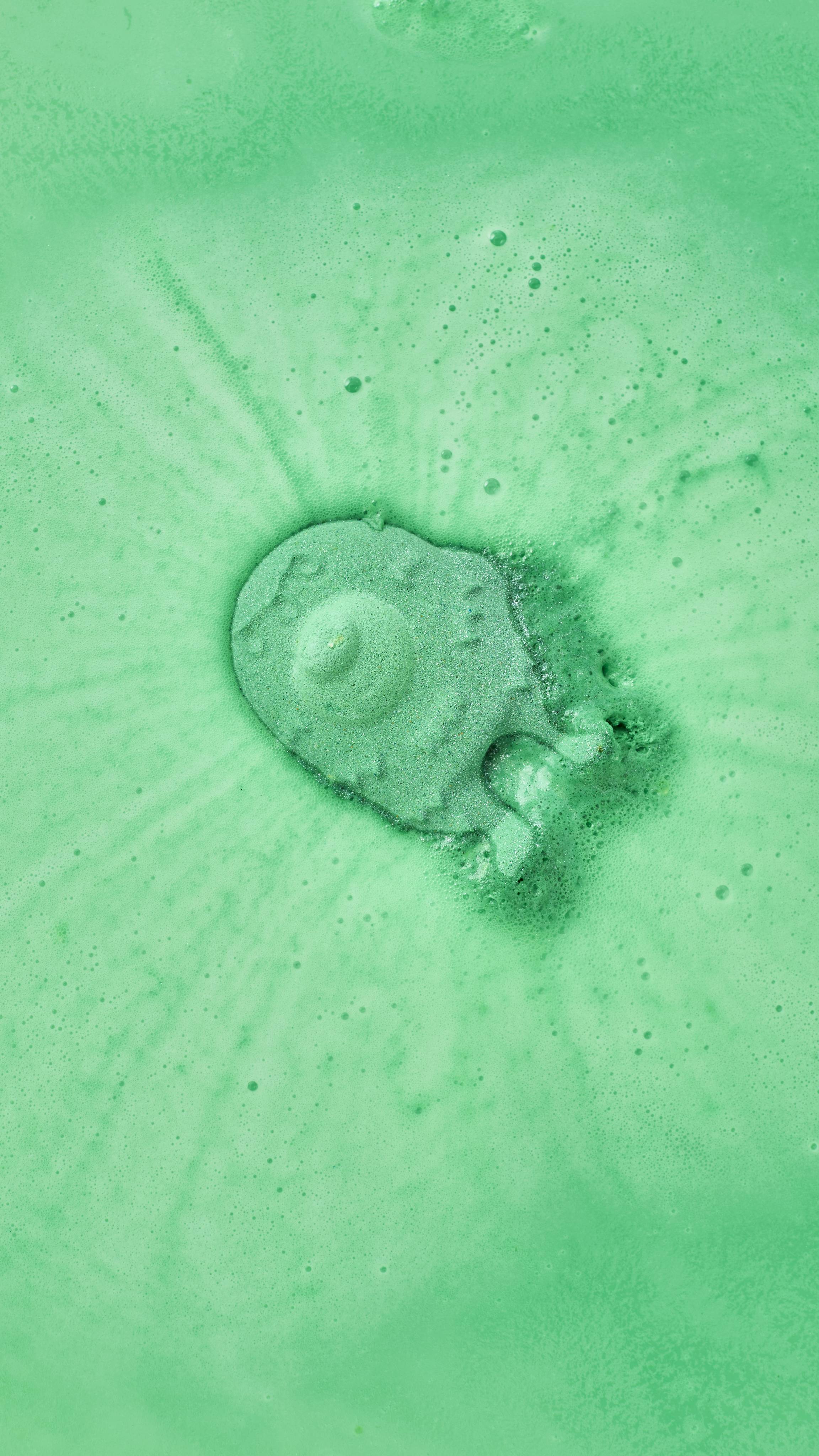 A mint-green, monster-shaped bath bomb dissolves at the centre of a bath, generating concentric ripples of lime-green and pale-green foam. The surrounding water is a vivid, opaque shade of green.