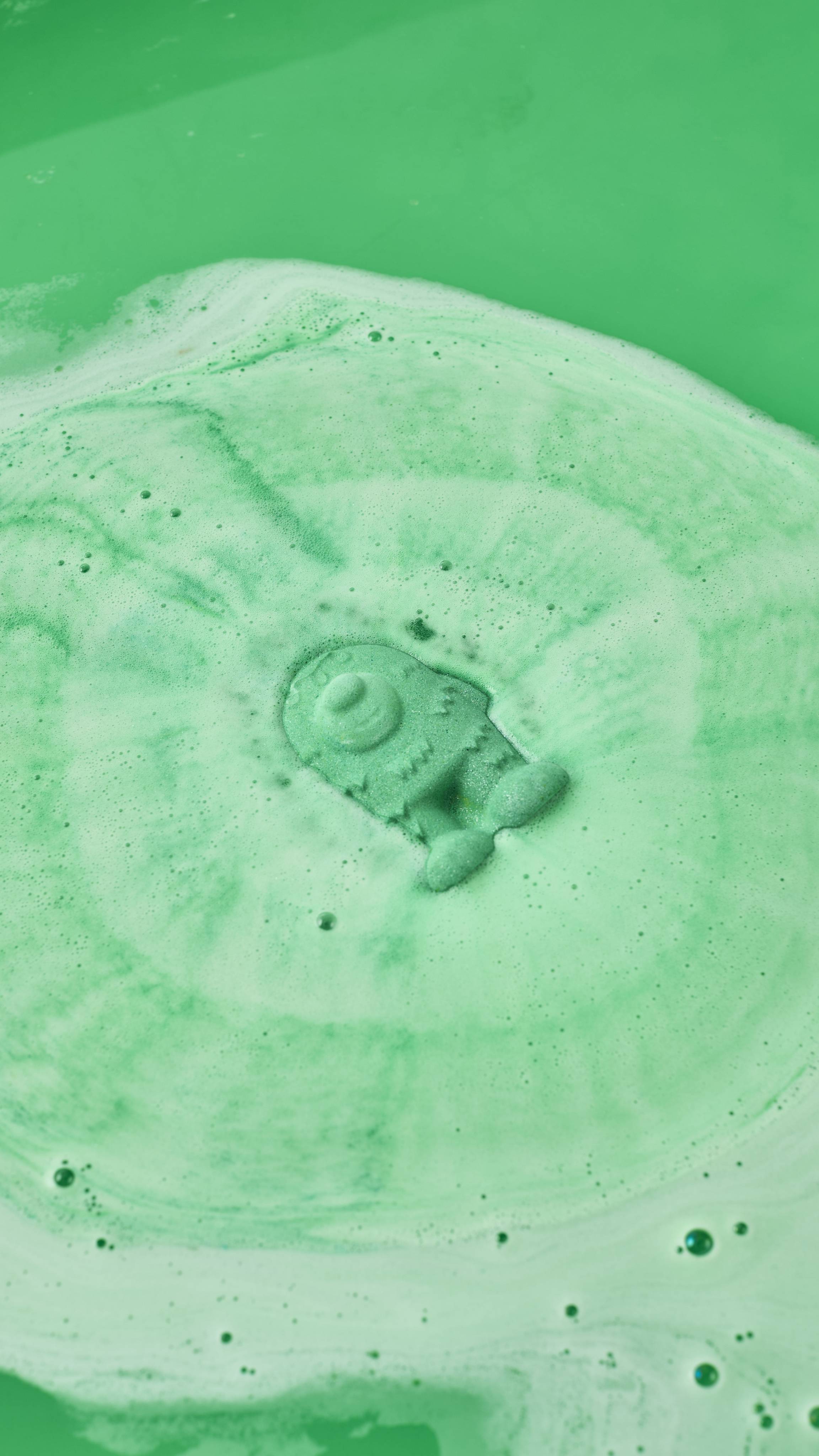 A mint-green, monster-shaped bath bomb dissolves at the centre of a bath, generating concentric ripples of lime-green and pale-green foam. The surrounding water is a vivid, opaque shade of green.