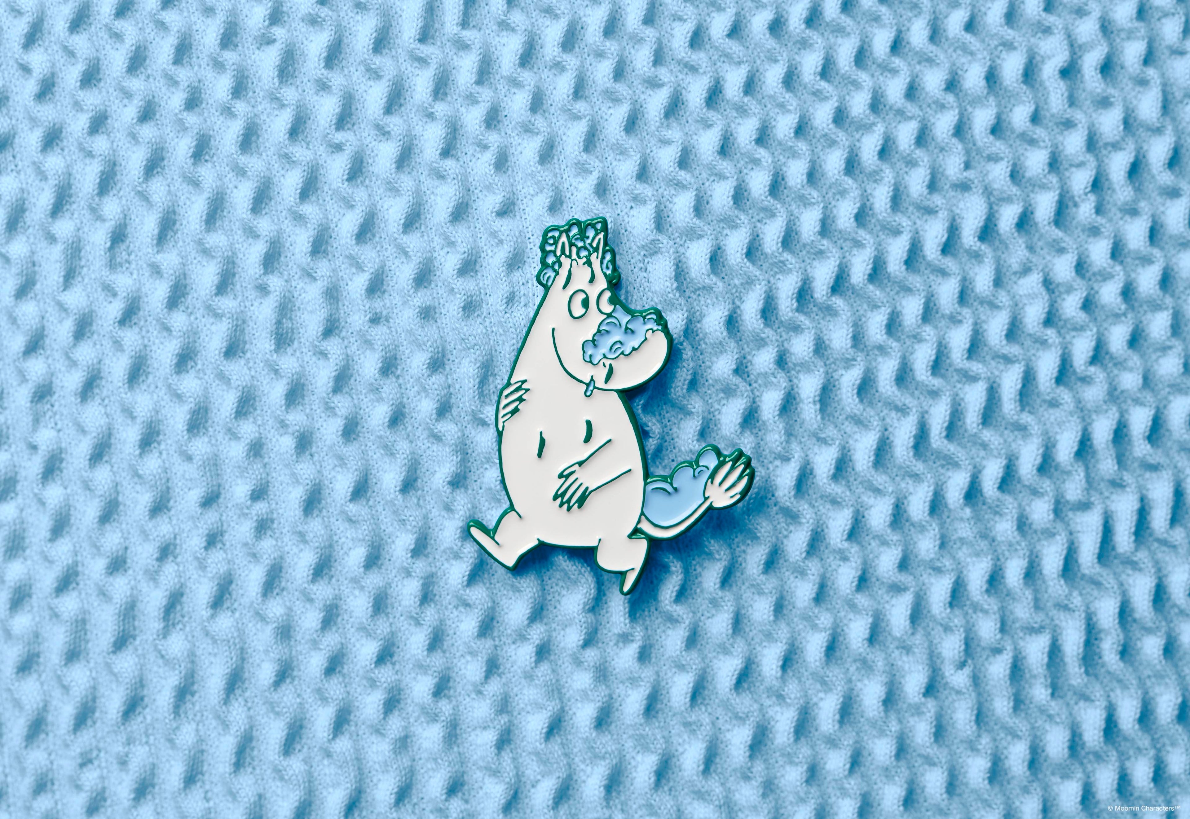 A Moomintroll enamel pin badge, showing the white character covered in blue soap suds on his nose, head, and tail. The pin is displayed in the centre of a textured, light blue waffle-weave fabric background.