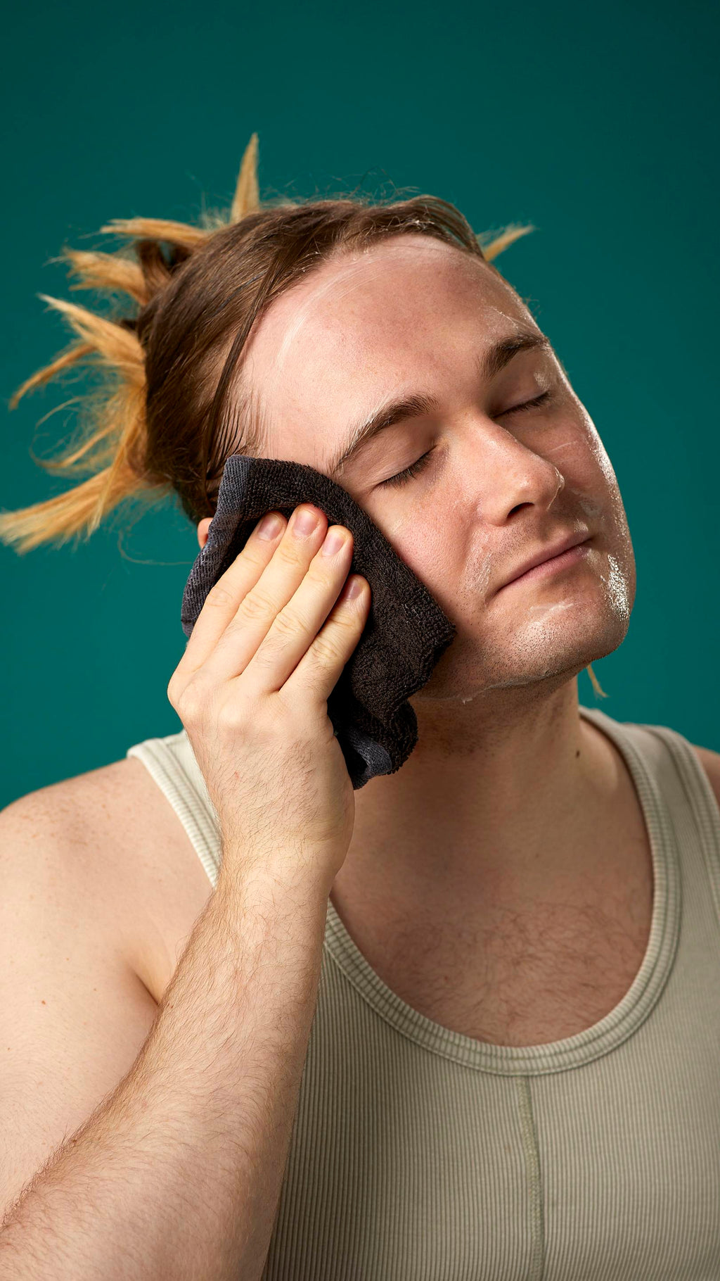 The image shows the model on a teal background with their hair tied back. They have a clean, fresh face as they gently remove the facial wash with a warm, damp flannel.