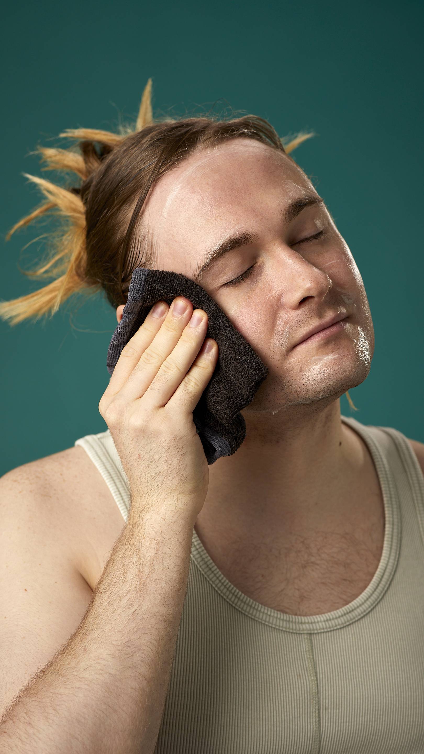 The image shows the model on a teal background with their hair tied back. They have a clean, fresh face as they gently remove the facial wash with a warm, damp flannel.