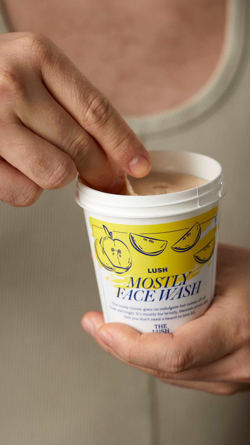 A close-up image of the model's hands. One hand holding the white, LUSH version-themed packed pot of the Mostly facial wash and scooping out the product with the other hand.