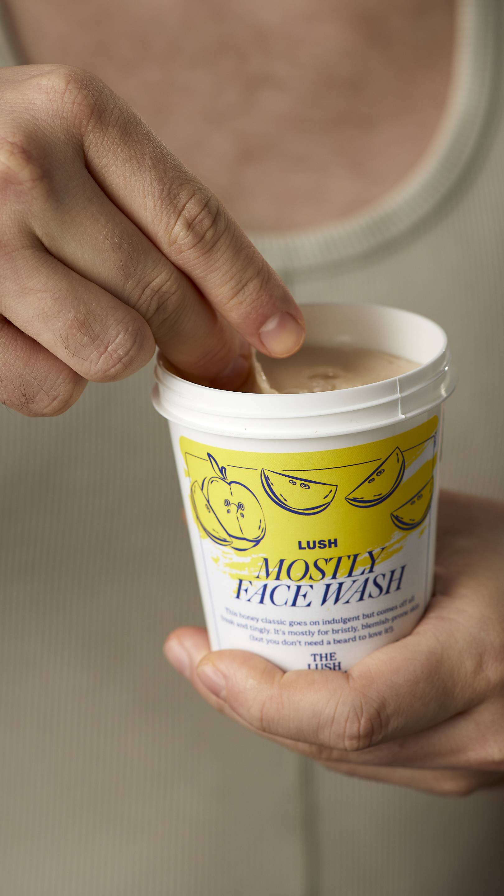 A close-up image of the model's hands. One hand holding the white, LUSH version-themed packed pot of the Mostly facial wash and scooping out the product with the other hand.