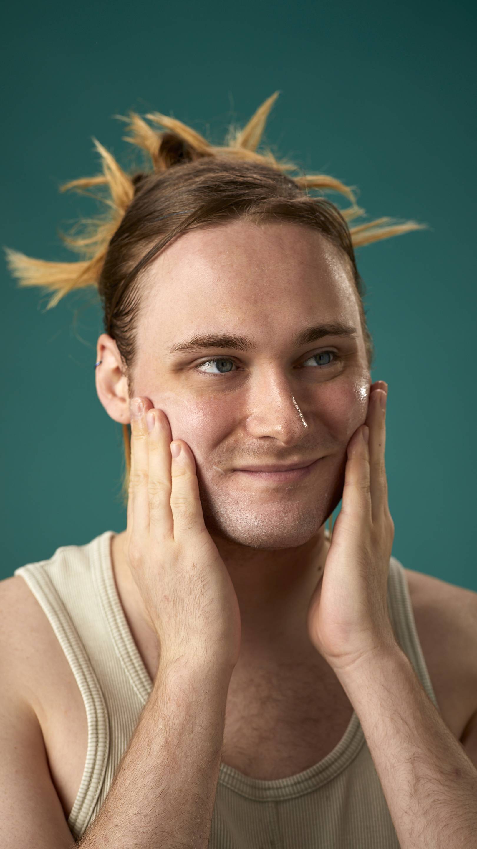 The image shows the model on a teal background with their hair tied back. They have a clean, fresh face as they gently apply the Mostly facial wash over their cheeks.