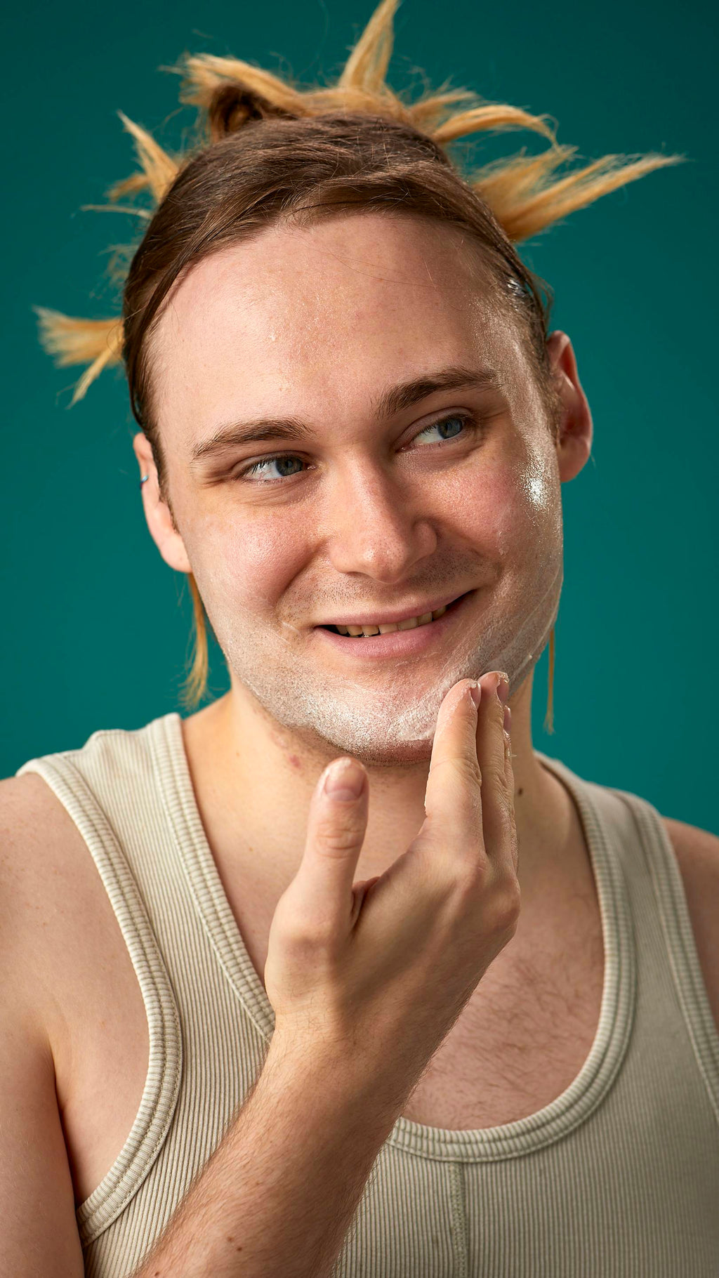 The image shows the model on a teal background with their hair tied back. They have a clean, fresh face as they gently apply the Mostly facial wash over their cheeks.