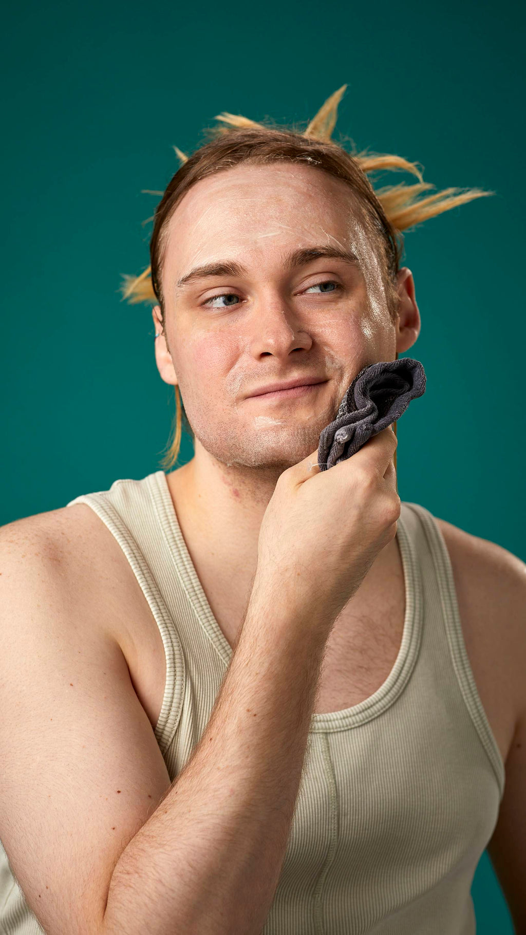 The image shows the model on a teal background with their hair tied back. They have a clean, fresh face as they gently remove the facial wash with a warm, damp flannel.