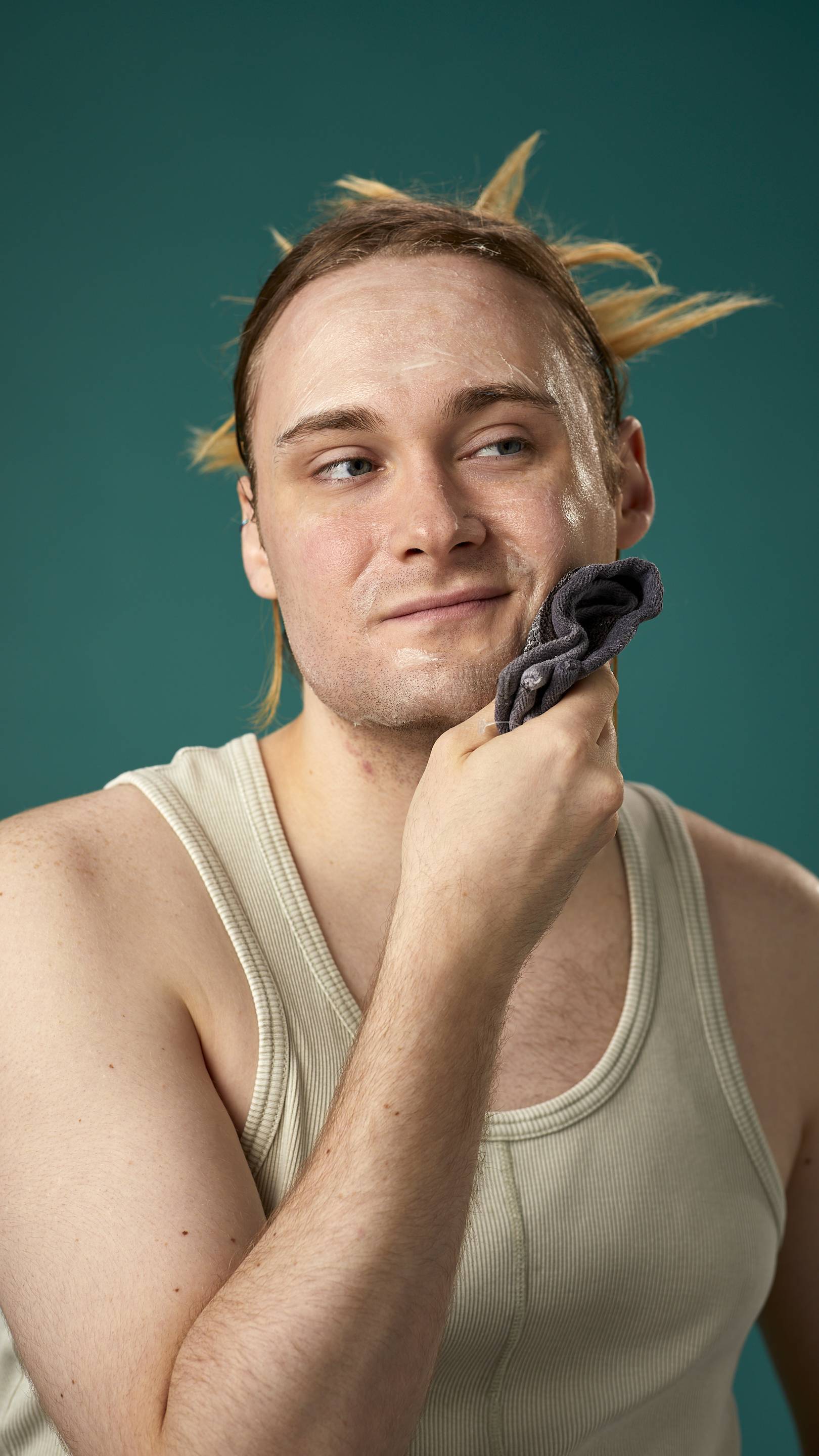 The image shows the model on a teal background with their hair tied back. They have a clean, fresh face as they gently remove the facial wash with a warm, damp flannel.