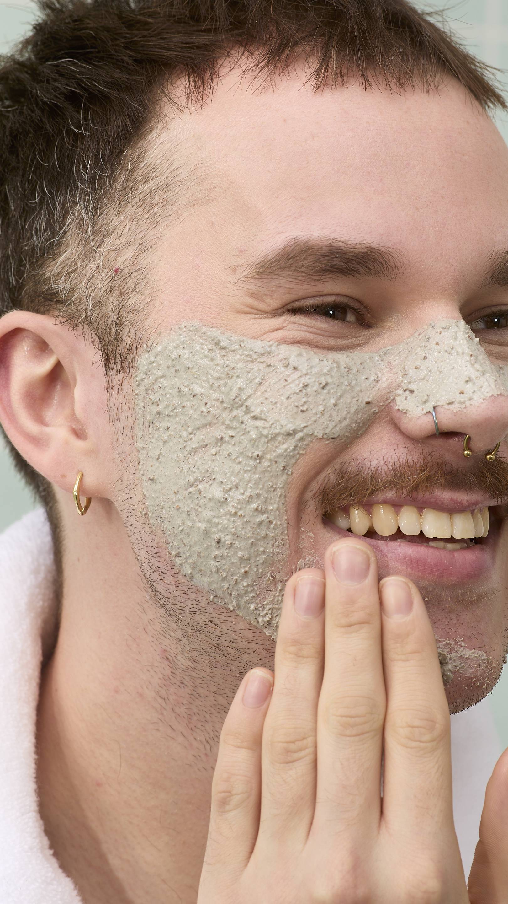 The image shows a model with a moustache and nose piercings smiling while applying a thick, grainy grey "Movis" face mask to their cheek and nose. They are wearing a white fluffy dressing gown against a background of pale green tiles.