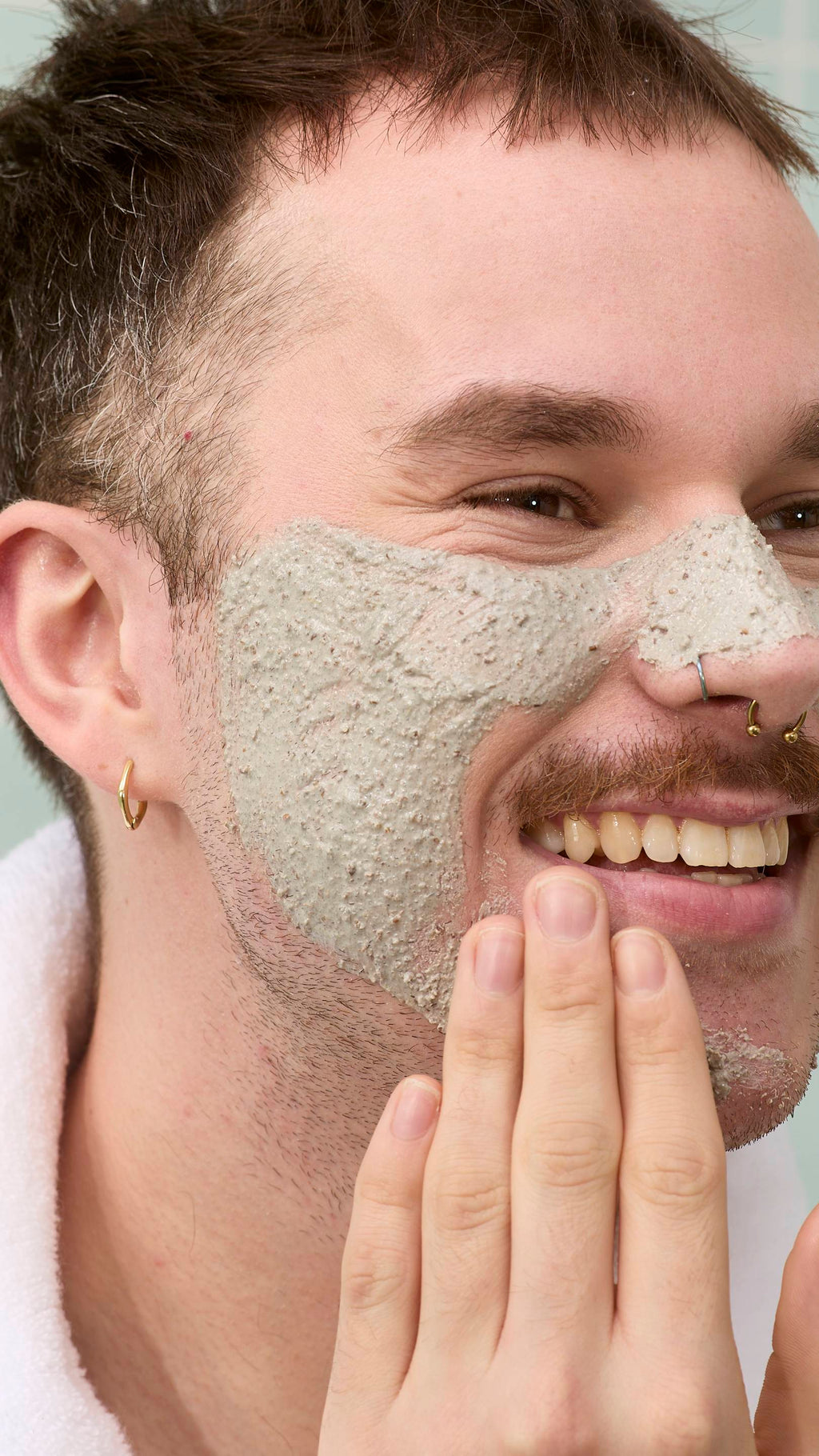 The image shows a model with a moustache and nose piercings smiling while applying a thick, grainy grey "Movis" face mask to their cheek and nose. They are wearing a white fluffy dressing gown against a background of pale green tiles.