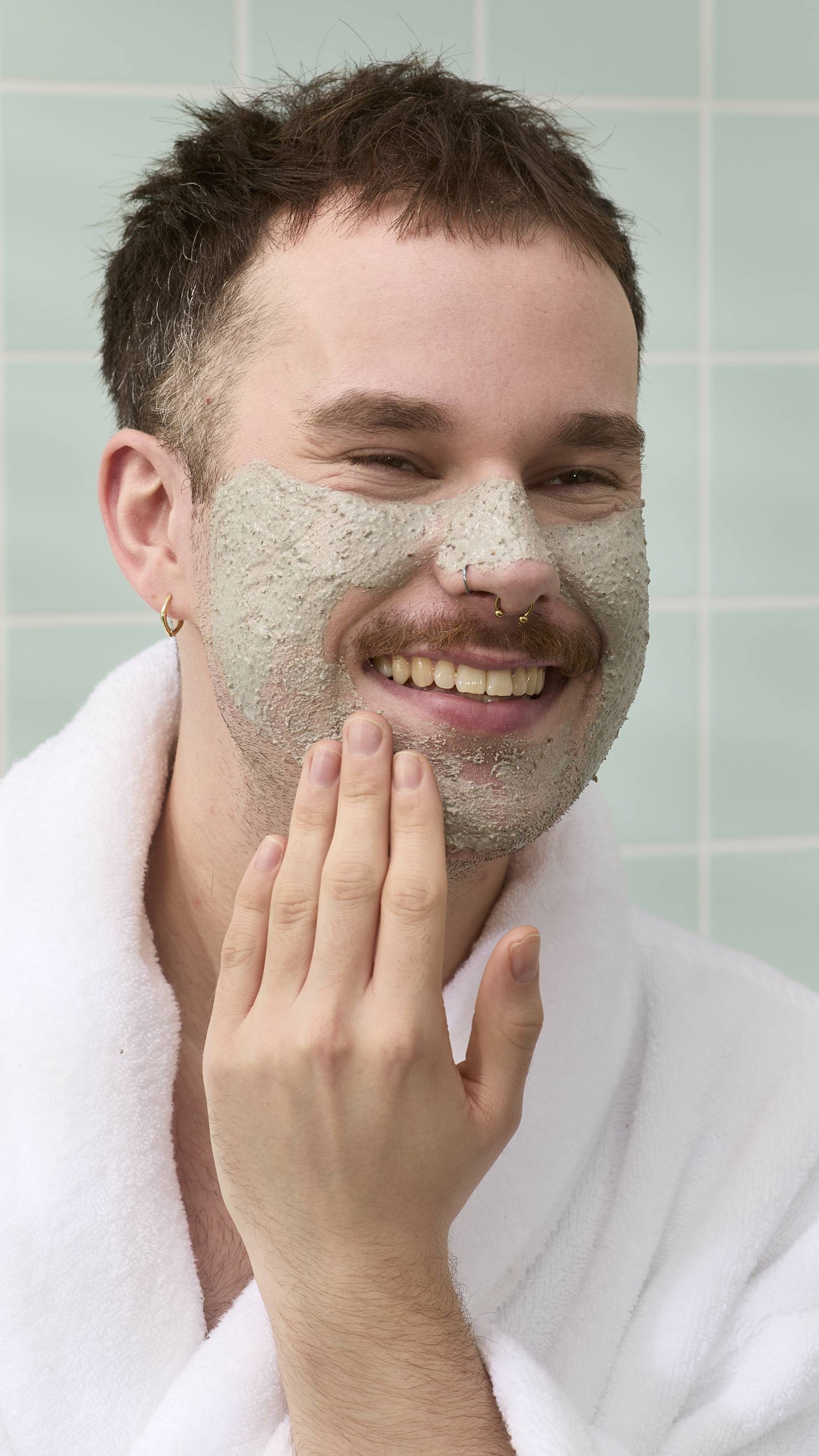 A model with a moustache and nose piercings is smiling while applying a textured grey "Movis" face mask. They are wearing a white bathrobe, set against a background of light green bathroom tiles.