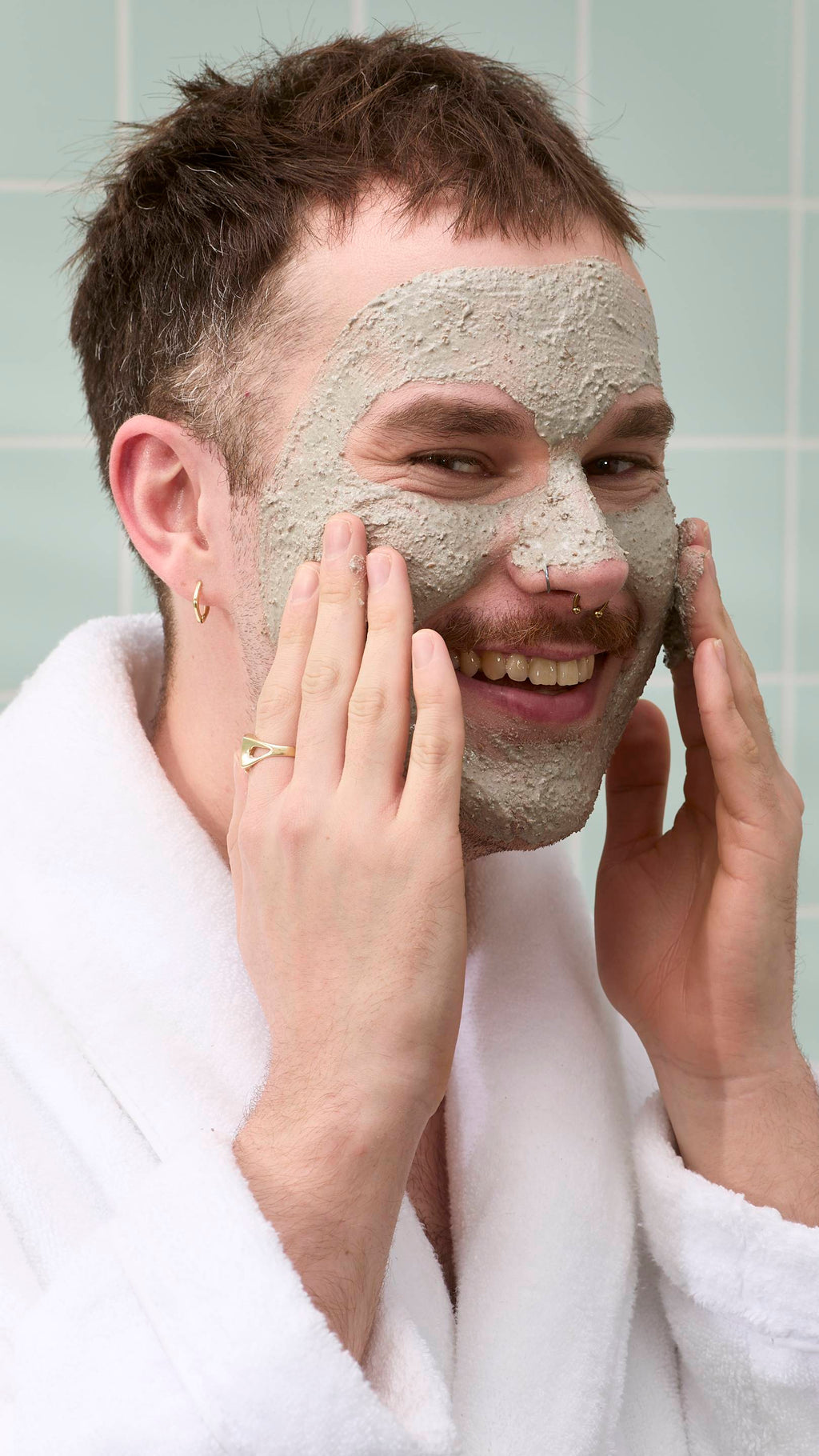 A close-up of a model with a moustache and nose piercings smiling while applying a textured grey "Movis" face mask with both hands. They are wearing a white dressing gown set against a background of light green bathroom tiles.