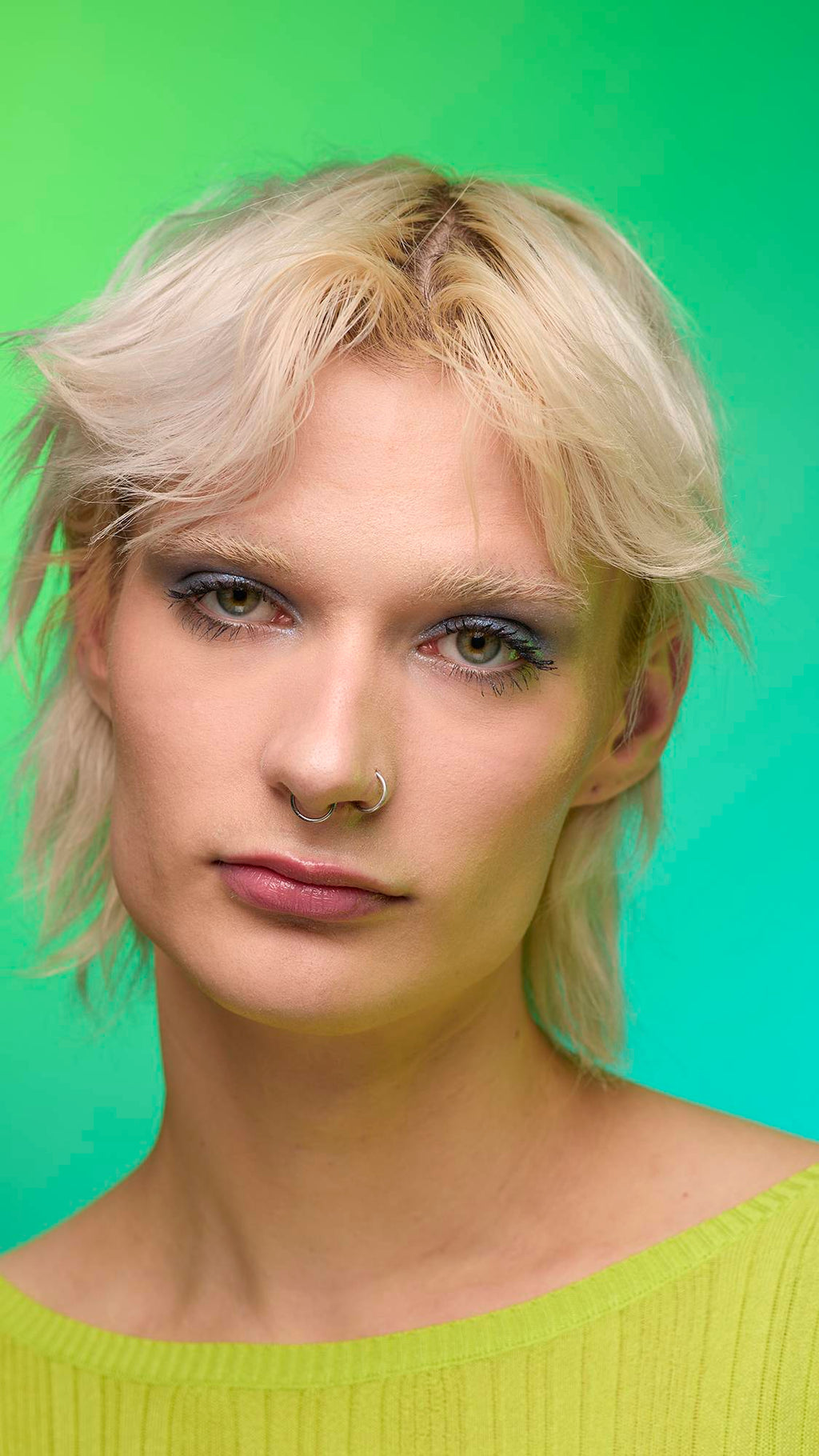The image shows a close-up of the model's face on a pale blue-to-green gradient background. The model's make-up is done with the focal point being a subtle, midnight-blue, shimmering eyeshadow made by the Nebula Glitter Crumble.