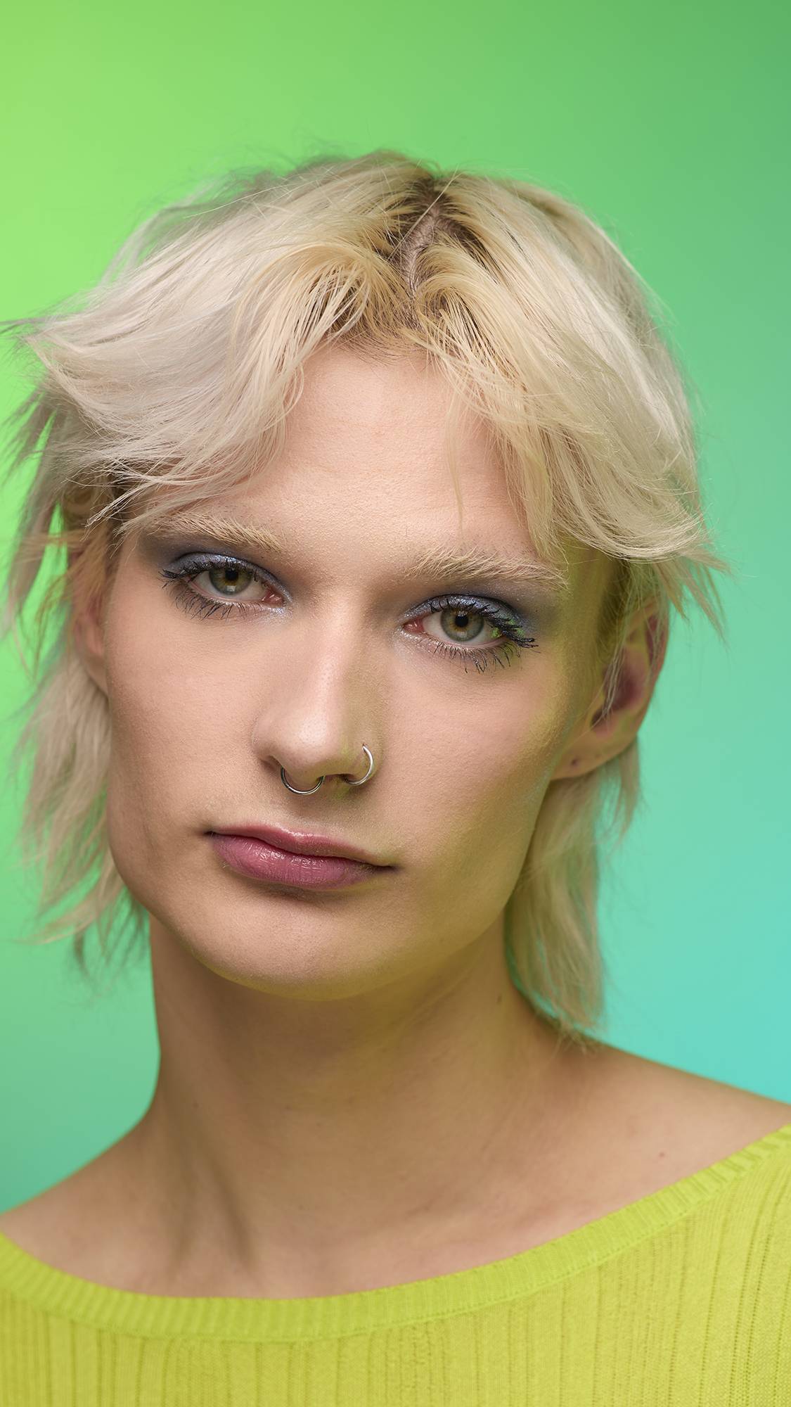The image shows a close-up of the model's face on a pale blue-to-green gradient background. The model's make-up is done with the focal point being a subtle, midnight-blue, shimmering eyeshadow made by the Nebula Glitter Crumble.
