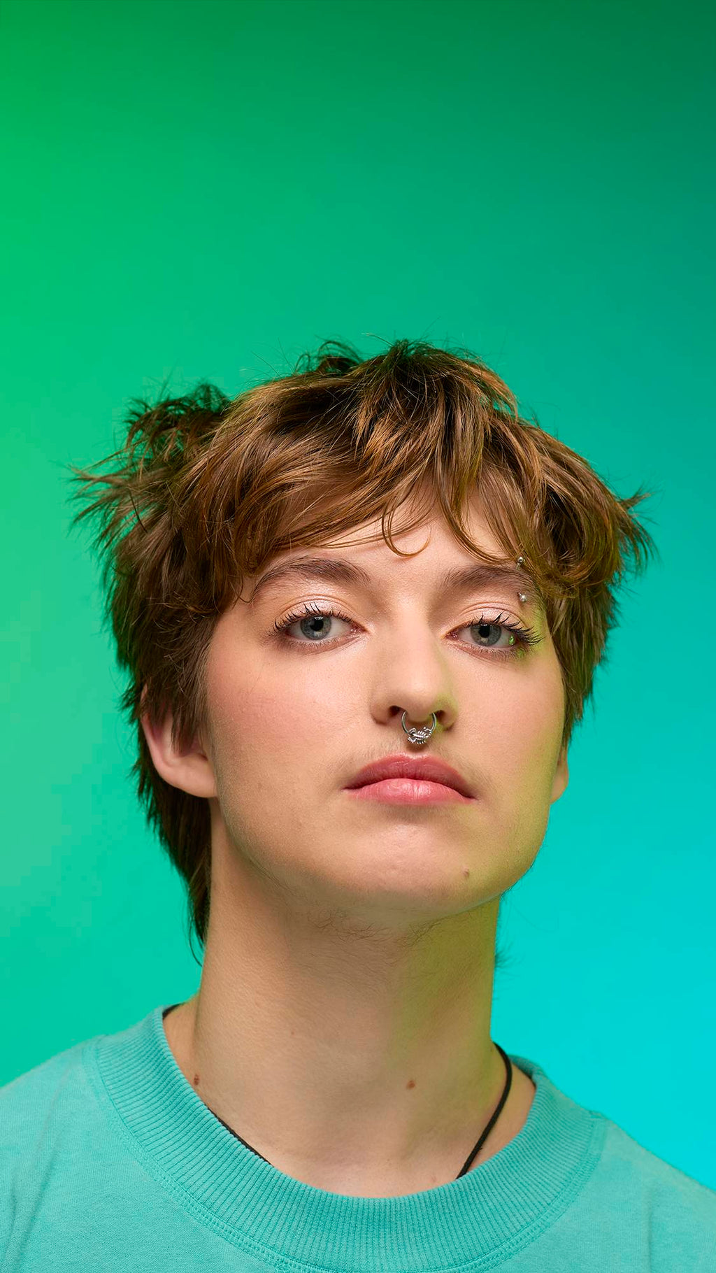 The image shows a close-up of the model's face on a pale blue-to-green gradient background. The model's make-up is done with the focal point being a subtle, iridescent, shimmering eyeshadow made by the New Moon Glitter Crumble.