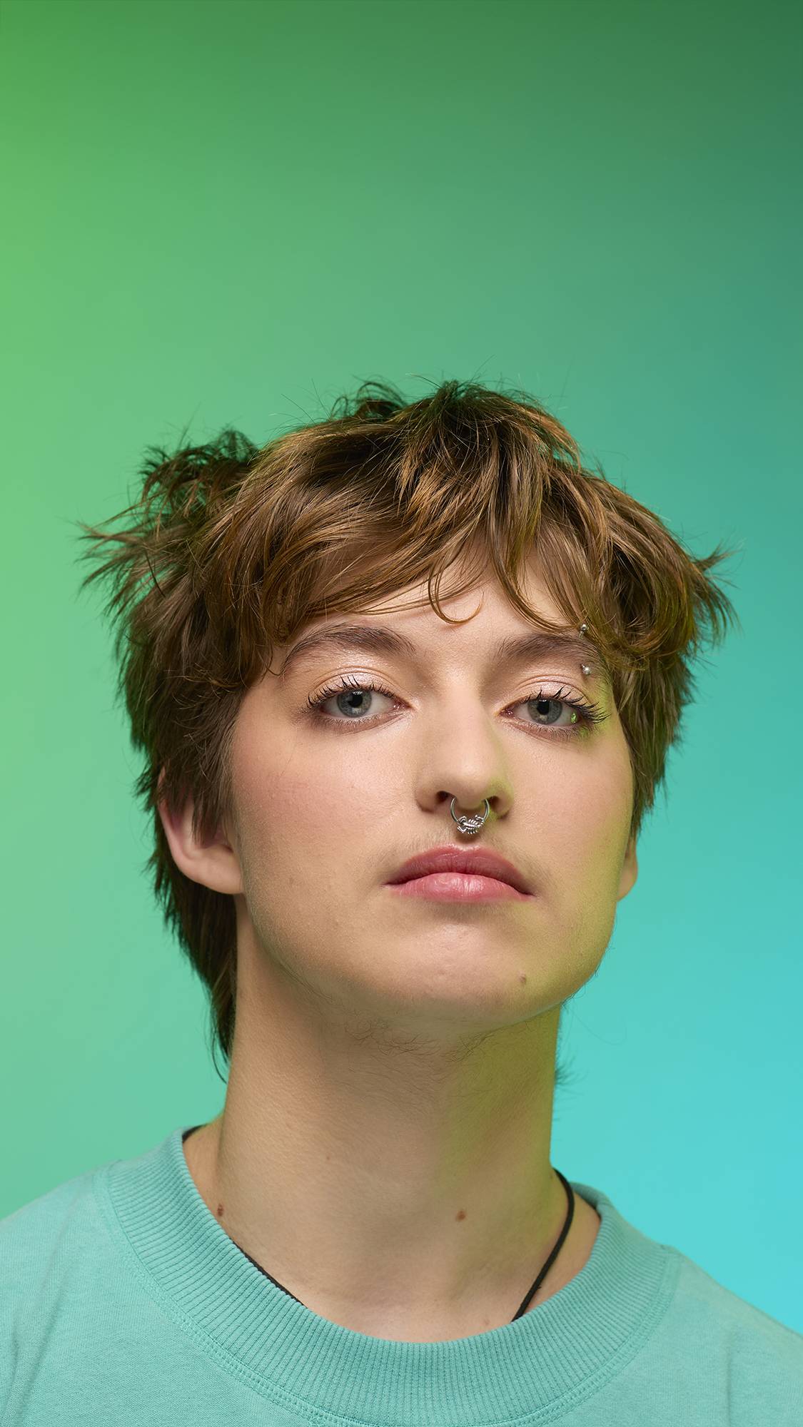 The image shows a close-up of the model's face on a pale blue-to-green gradient background. The model's make-up is done with the focal point being a subtle, iridescent, shimmering eyeshadow made by the New Moon Glitter Crumble.