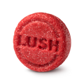 New shampoo. A circular, highly textured red solid shampoo bar. The fibrous, shredded surface features the brand name 'LUSH' embossed clearly in the centre of the round face.