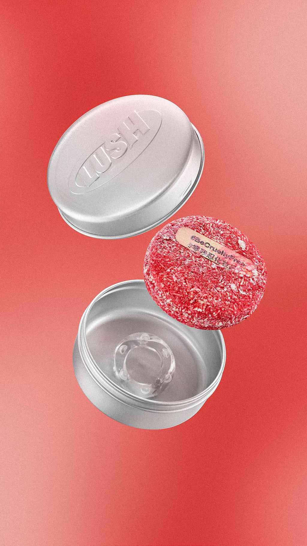 Floating against a warm red gradient, a textured red circular "NEW" shampoo bar with a small wooden tab is shown alongside an open, silver aluminium tin and its lid, which is embossed with the 'LUSH' logo.