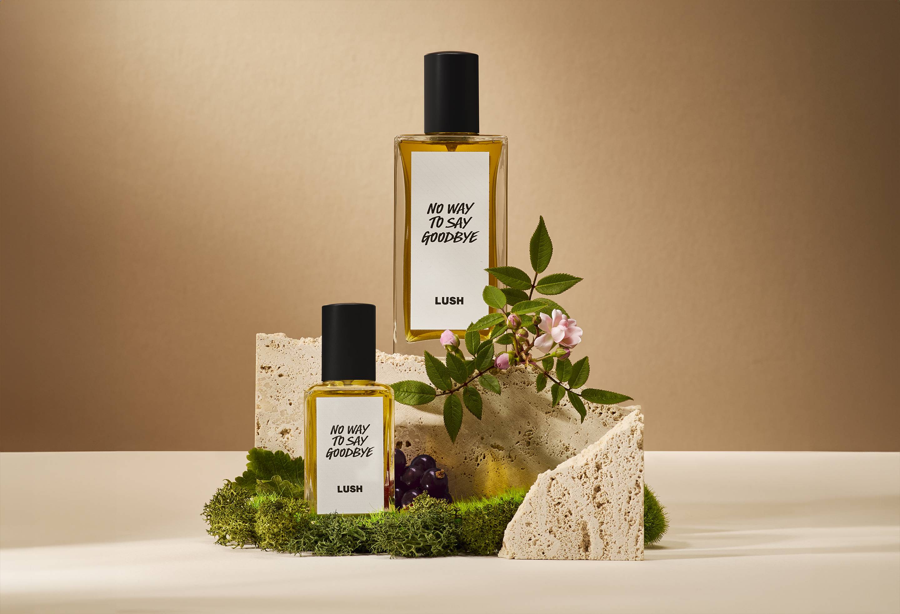 The image shows a soft, beige background. In the centre of focus are two bottles of two different sizes of perfume sitting on top of moss and rock with moss, blackcurrant and litsea cubeba flowers.