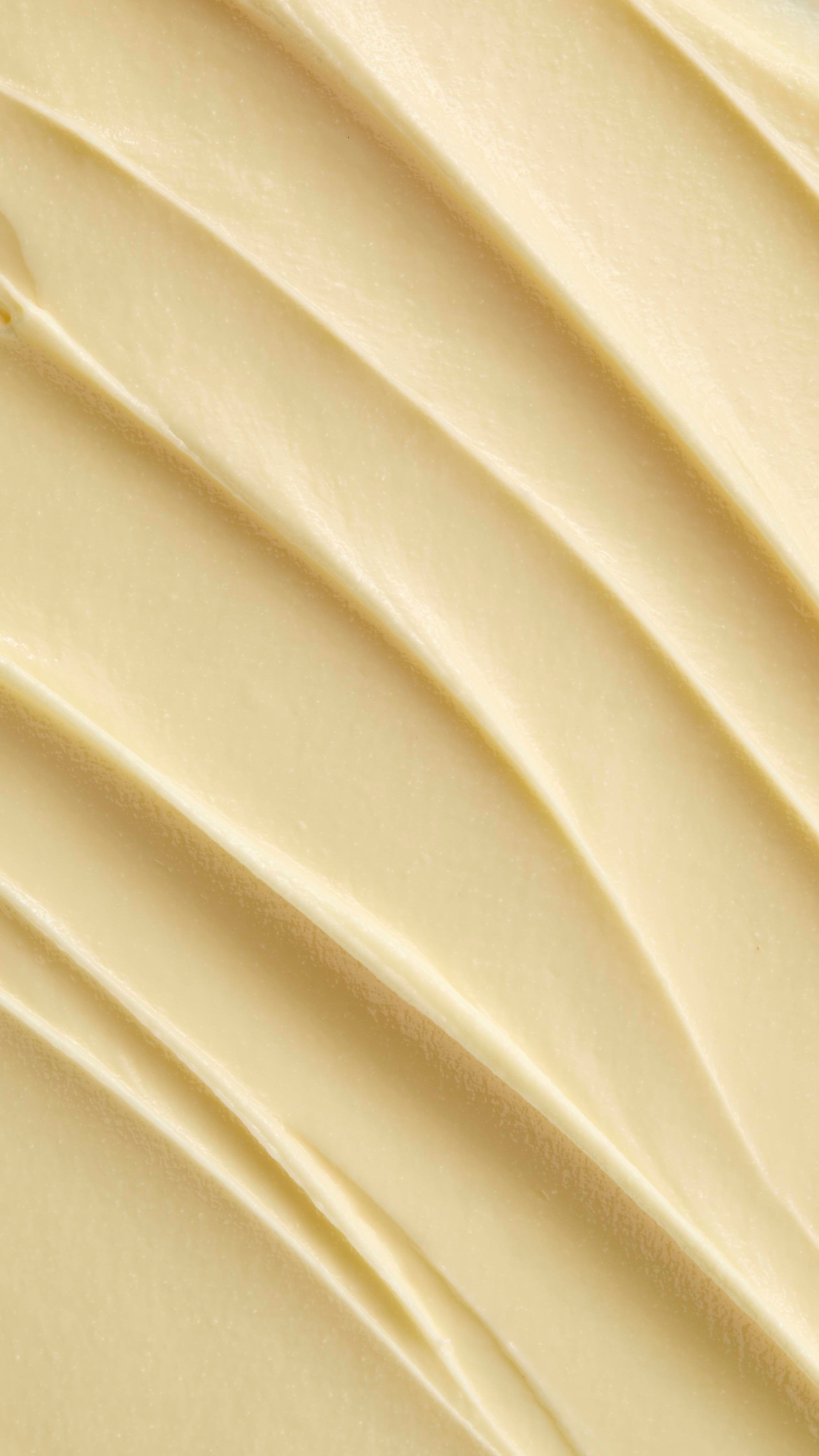 A super close-up image showing diagonal, overlapping smears of the thick, creamy, pale yellow Lush "Oat" facial cleanser. The smooth product has a matte finish and subtle ridged textures.