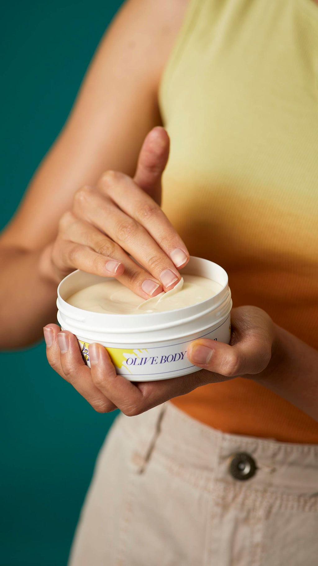 A close-up shot of the model's hands. One hand is holding the flat round, white, LUSH Version-themed packed tub of Olive body butter.