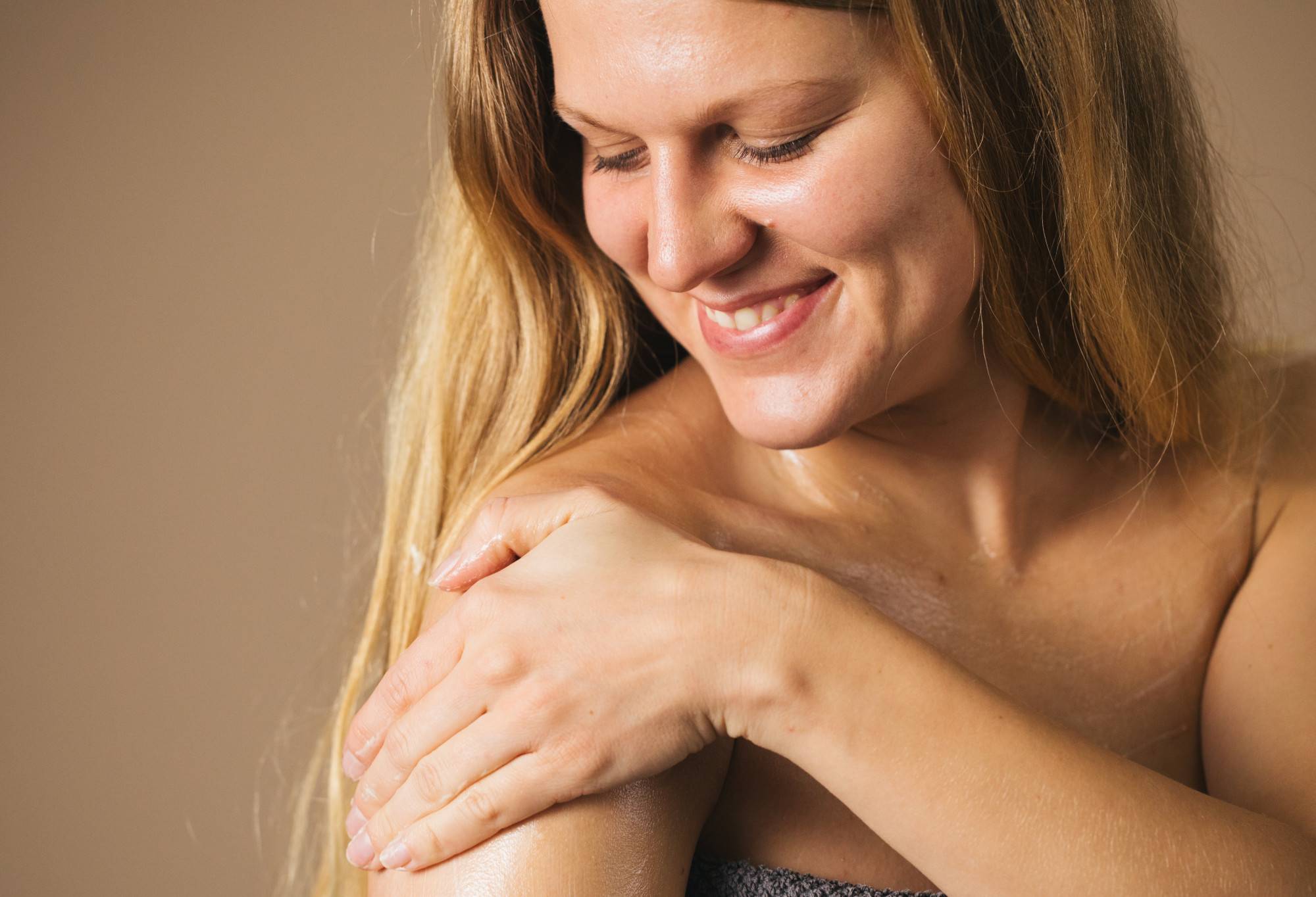 A person with long, blonde hair is smiling ear to ear as they look down and rub Pansy body lotion onto their shoulder.