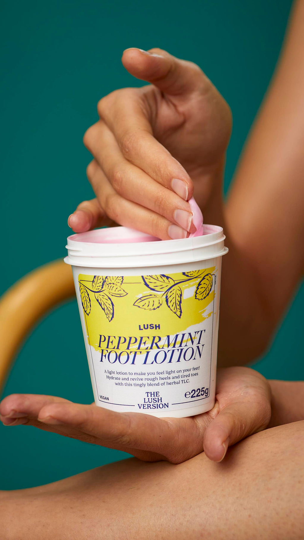 The image shows the model's arms stretched into the centre holding the white, LUSH Version-themed packed pot of the Peppermint foot lotion. One of their hands is scooping the product out. There is a teal background.