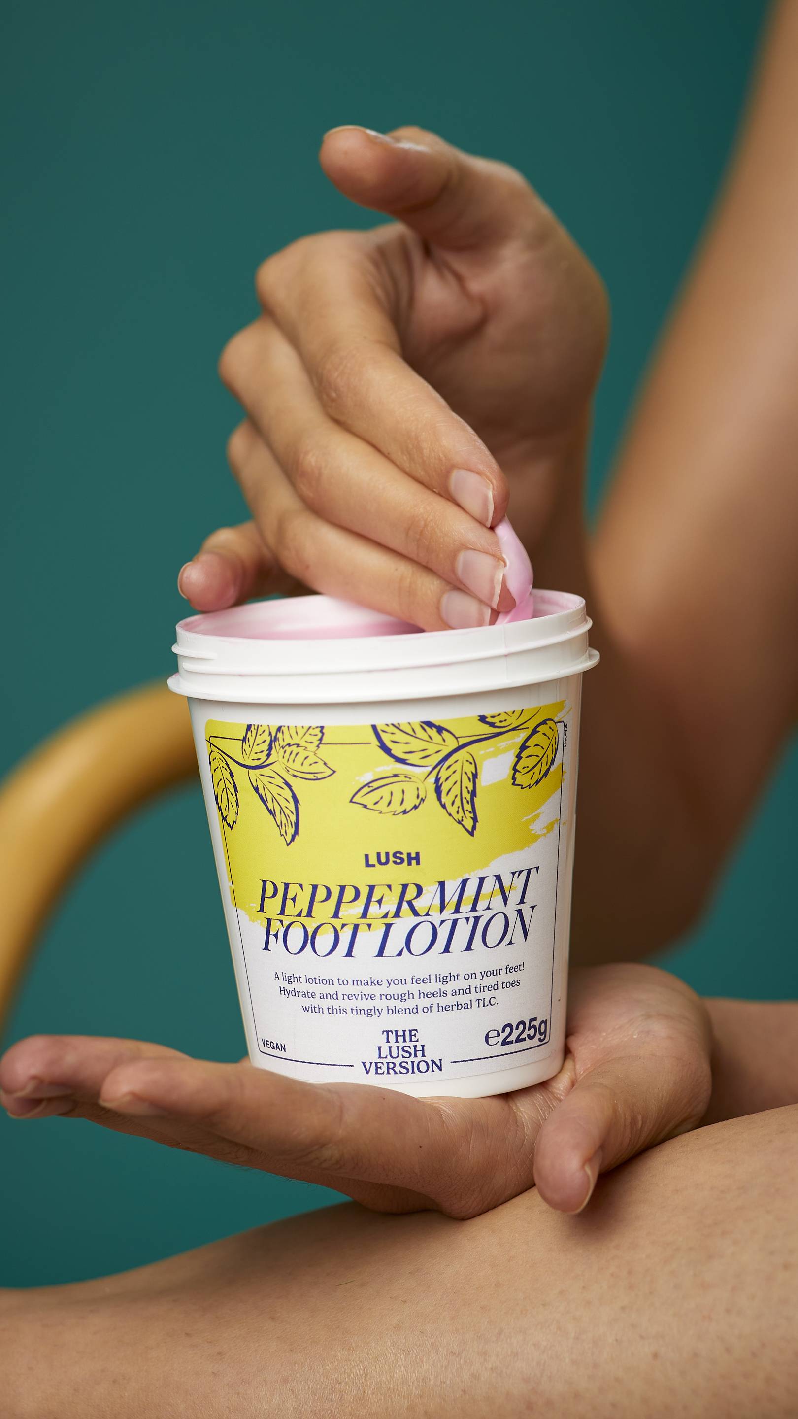 The image shows the model's arms stretched into the centre holding the white, LUSH Version-themed packed pot of the Peppermint foot lotion. One of their hands is scooping the product out. There is a teal background.