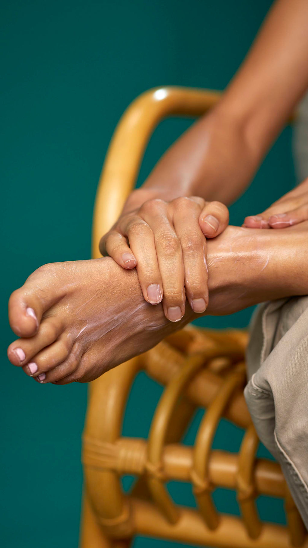 The image shows a close-up of the model's lower legs as they are sat on a wicker chair on a tea background. Their foot is resting on their leg and their toenails are painted pastel pink as they apply the Peppermint foot lotion.
