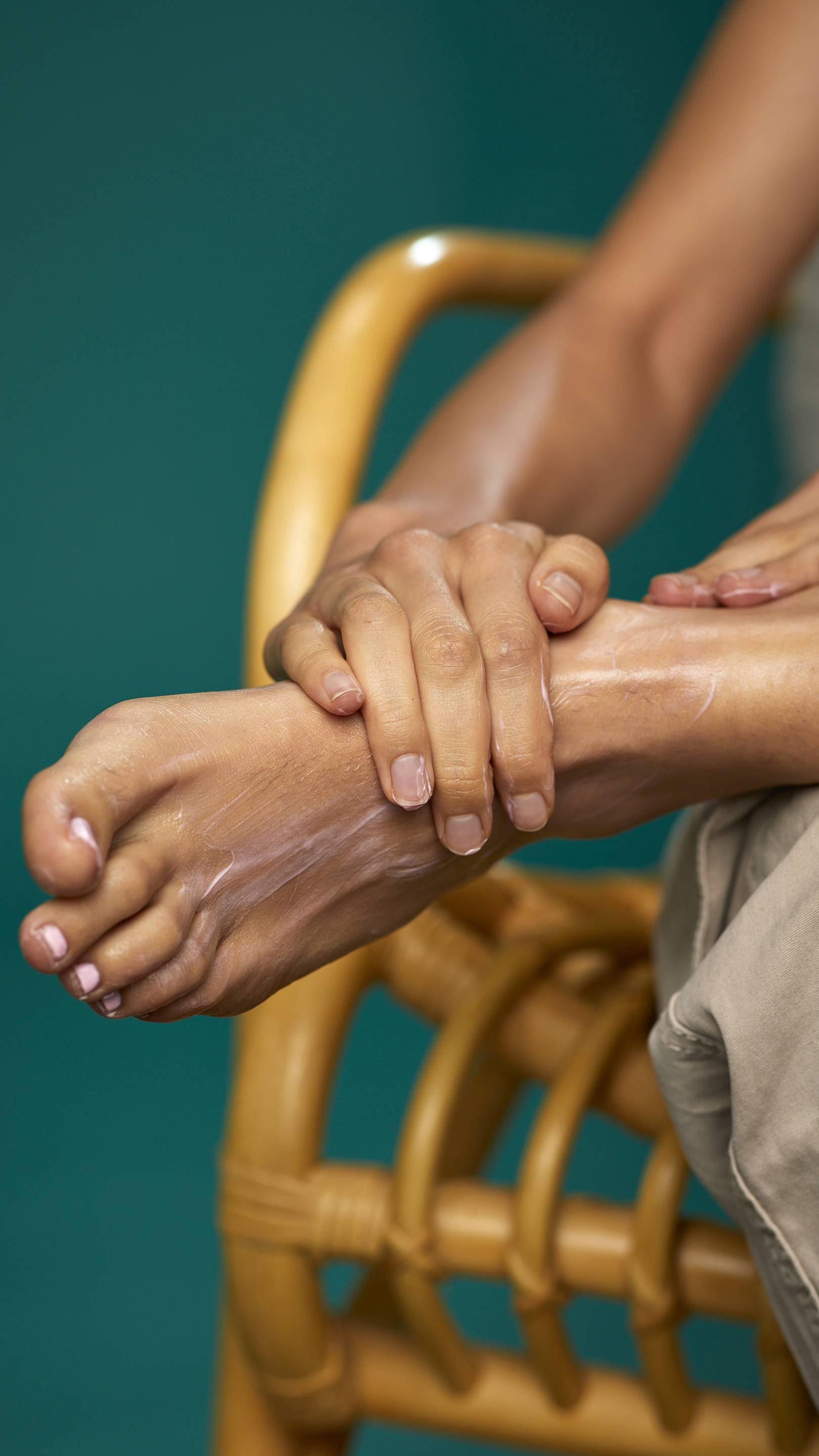The image shows a close-up of the model's lower legs as they are sat on a wicker chair on a tea background. Their foot is resting on their leg and their toenails are painted pastel pink as they apply the Peppermint foot lotion.
