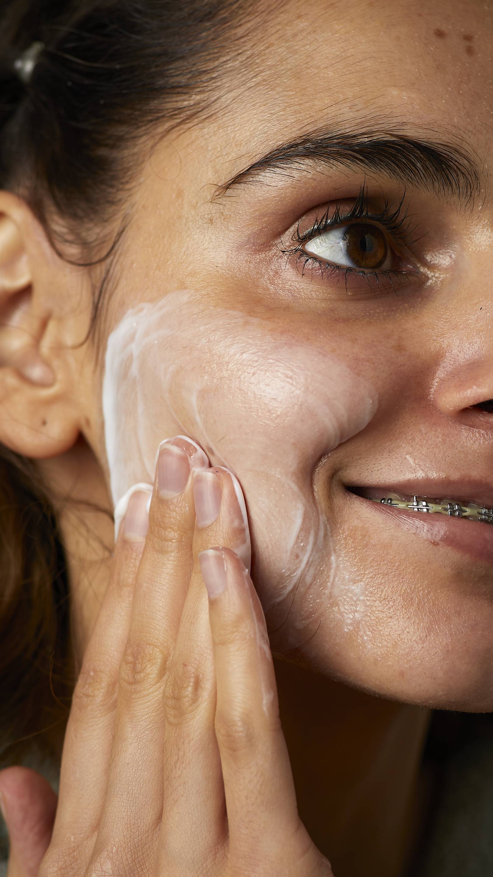 A super close-up image of one of the model's cheeks. They are smiling showing a glimpse of braces as they gently apply the Pineapple facial wash in circular motions over their cheek.