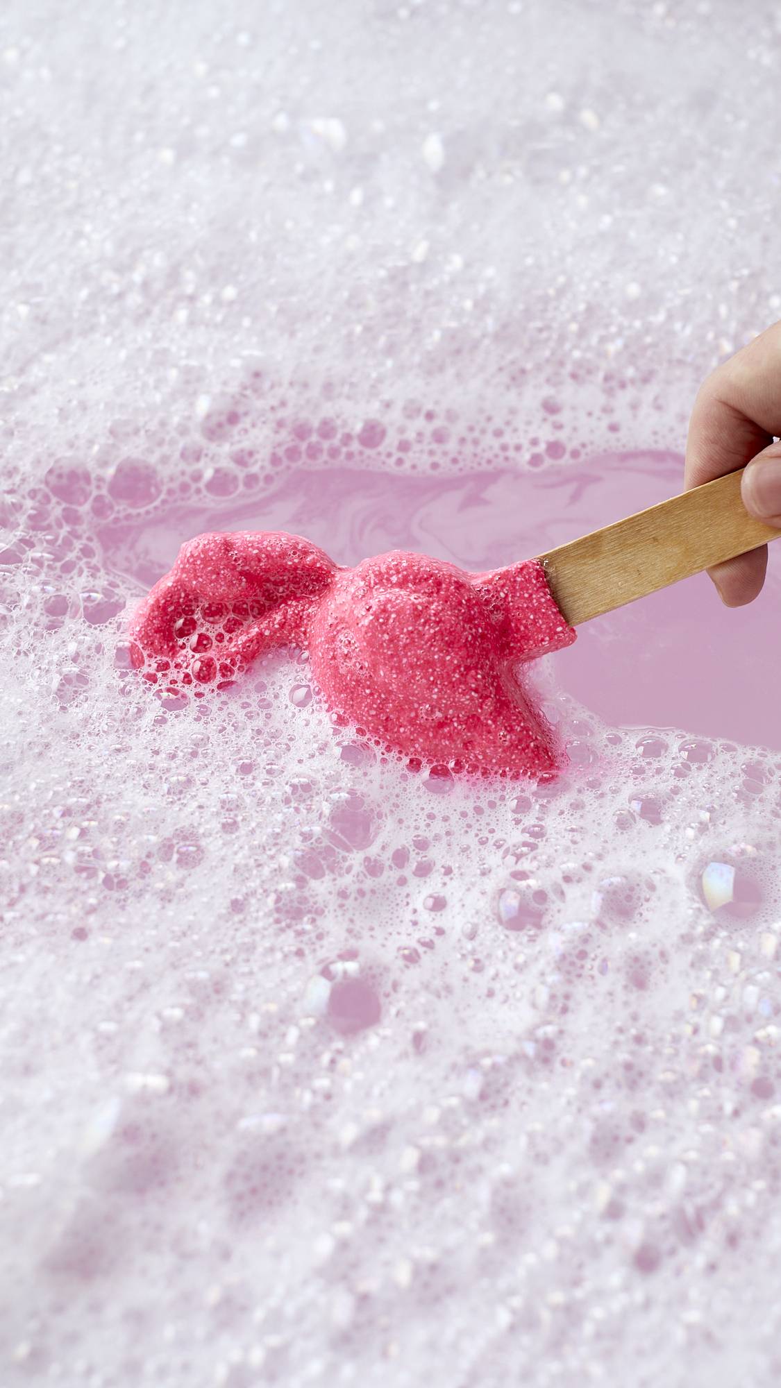 The image shows a close-up shot of the Pink Flamingo reusable bubble bar stick being swirled around delicate, pink bath water surrounded by a thick blanket of foamy bubbles made by the product.