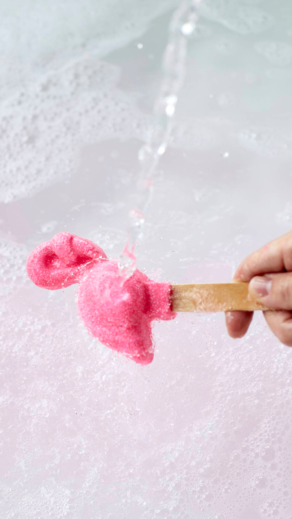 The image shows a close-up shot of the Pink Flamingo reusable bubble bar stick being held under running tap water. The Pink flamingo at the end creates bubbly, delicately rose-tinted water below.
