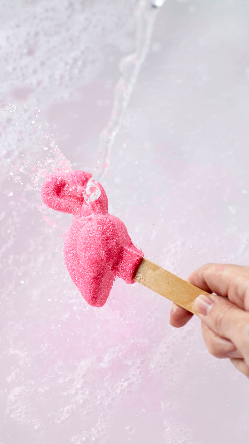 The image shows a close-up shot of the Pink Flamingo reusable bubble bar stick being held under running tap water. The Pink flamingo at the end creates bubbly, delicately rose-tinted water below.