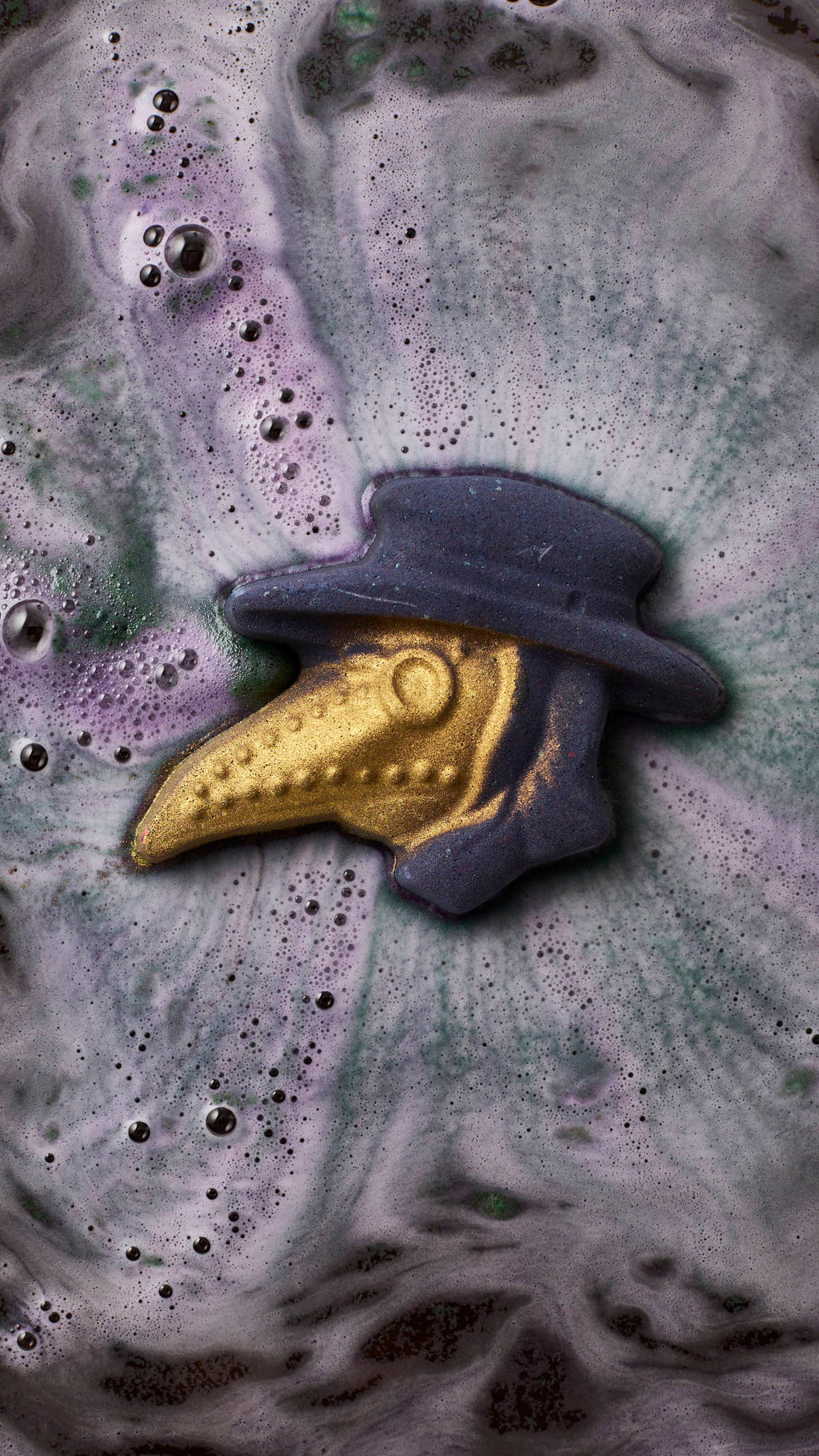 A plague doctor-shaped bath bomb dissolves in a bath, its dark purple hat and shimmering gold-beaked mask floating amidst swirling, bubbly trails of grey and violet foam.