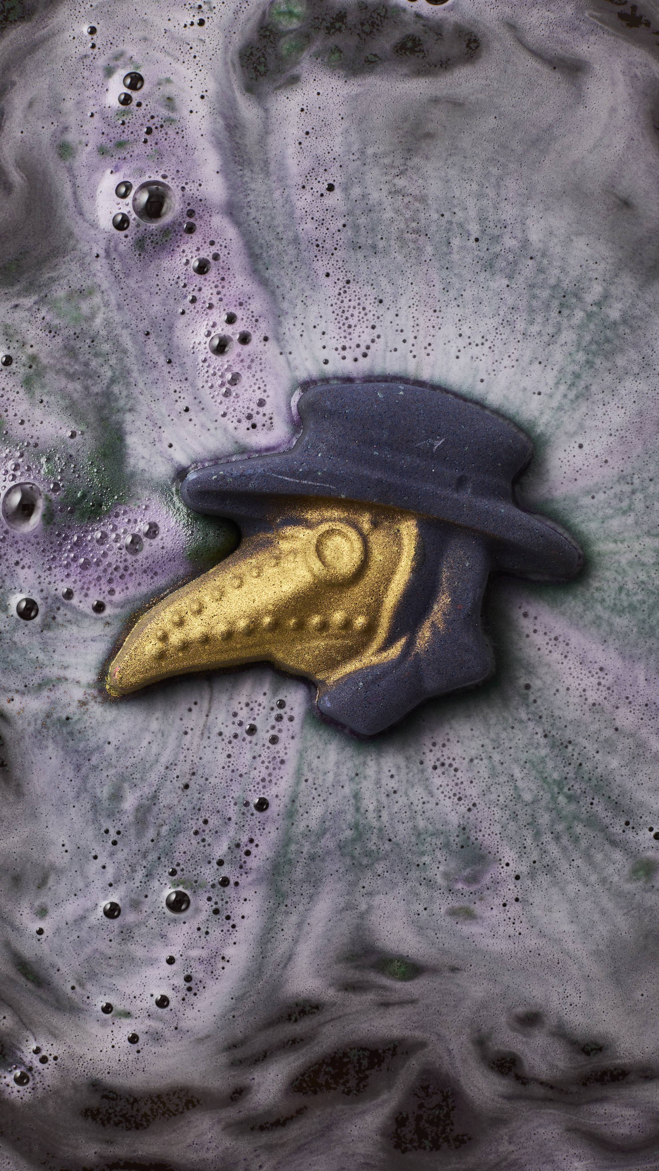 A plague doctor-shaped bath bomb dissolves in a bath, its dark purple hat and shimmering gold-beaked mask floating amidst swirling, bubbly trails of grey and violet foam.