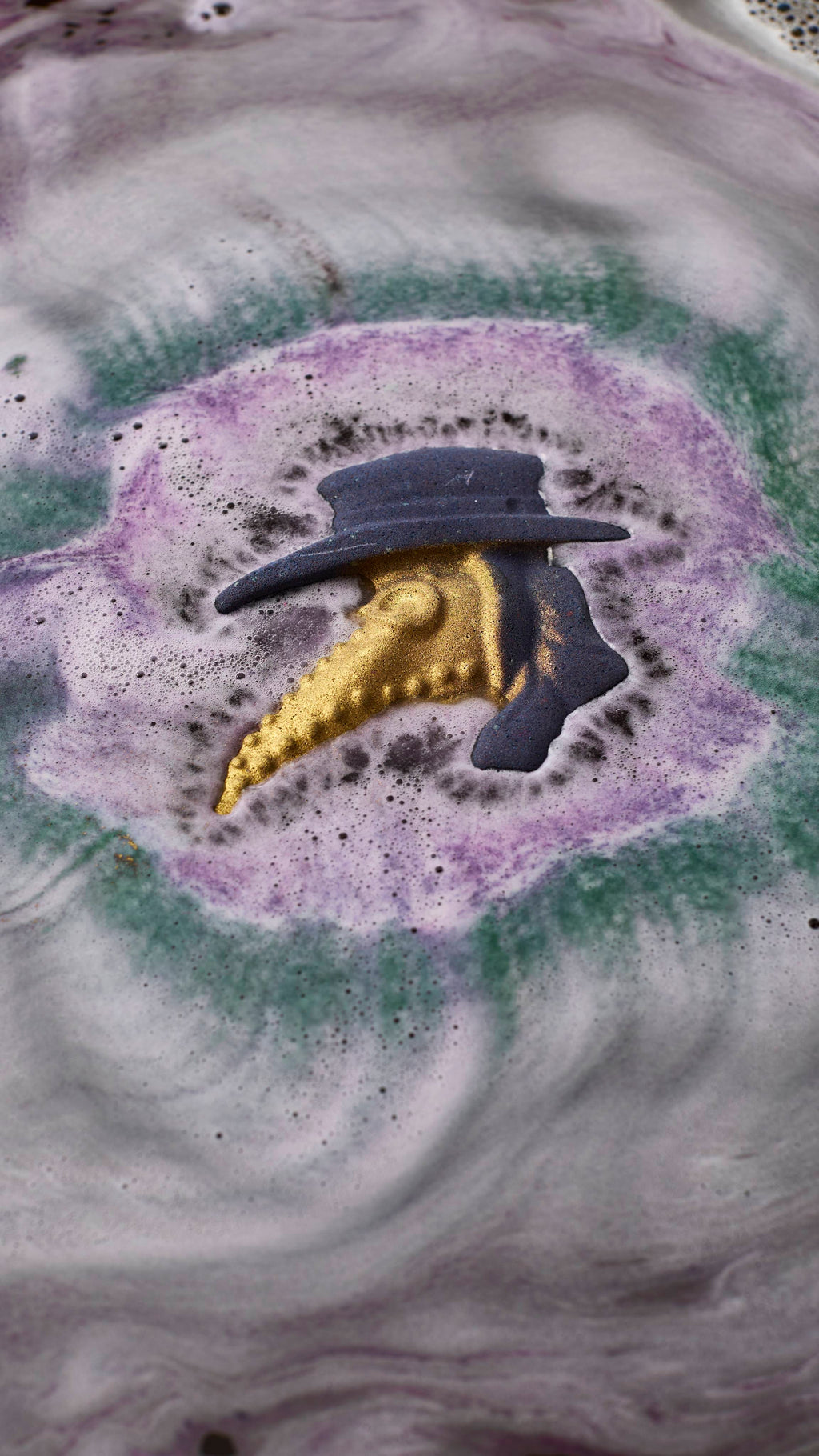A plague doctor-shaped bath bomb dissolves in water, creating concentric ripples of purple, green, and dark grey froth around its purple hat and shimmering gold-beaked mask. Swirling patterns of light grey foam cover the rest of the bath.