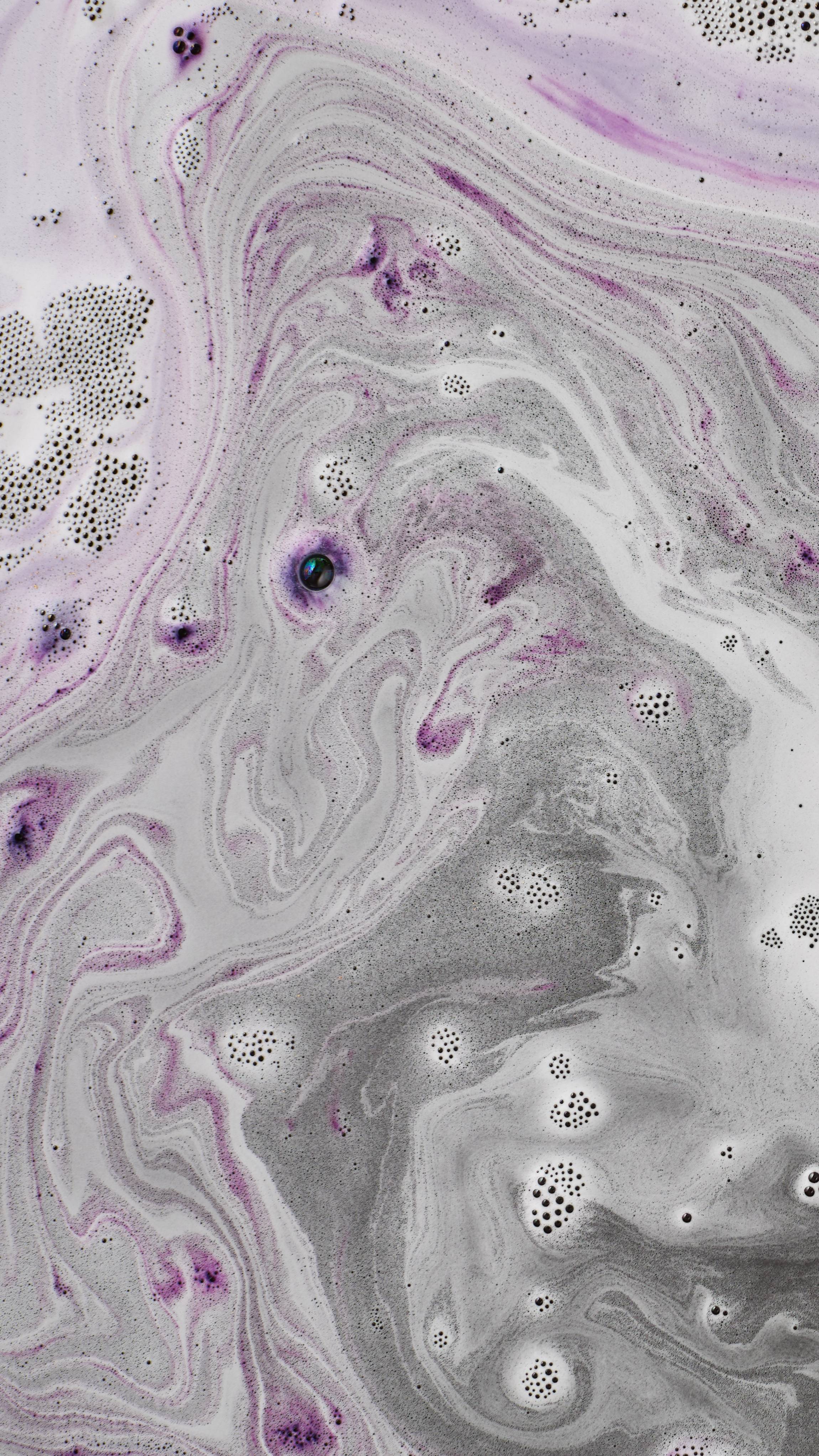 An abstract aerial shot of bath bomb foam swirling in water, creating intricate, marbled waves of dark grey, pale white, and streaks of violet. Fine, clusters of dark soapy bubbles are scattered across the textured, frothy surface.