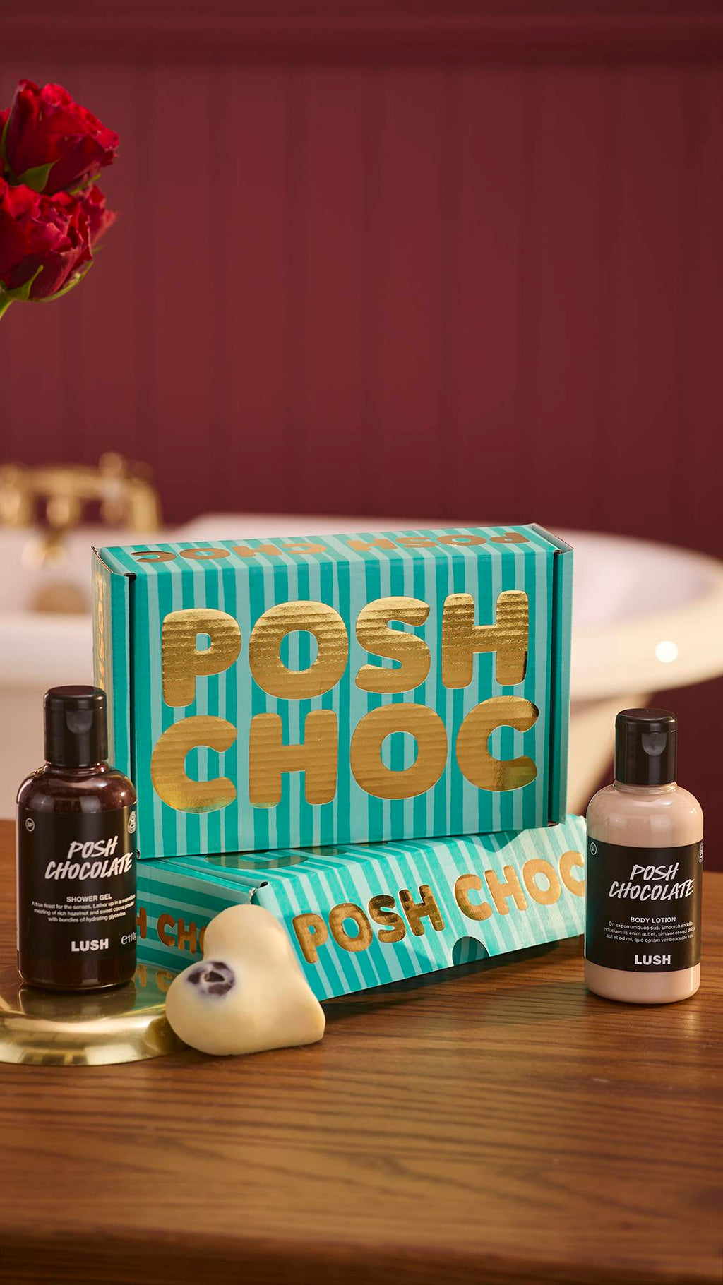 The image shows a dark, chocolate-coloured background with roses just visible on the left. Two stacked Posh Choc, teal-striped gift boxes sit on a wooden counter with the three, gourmand included products around them.