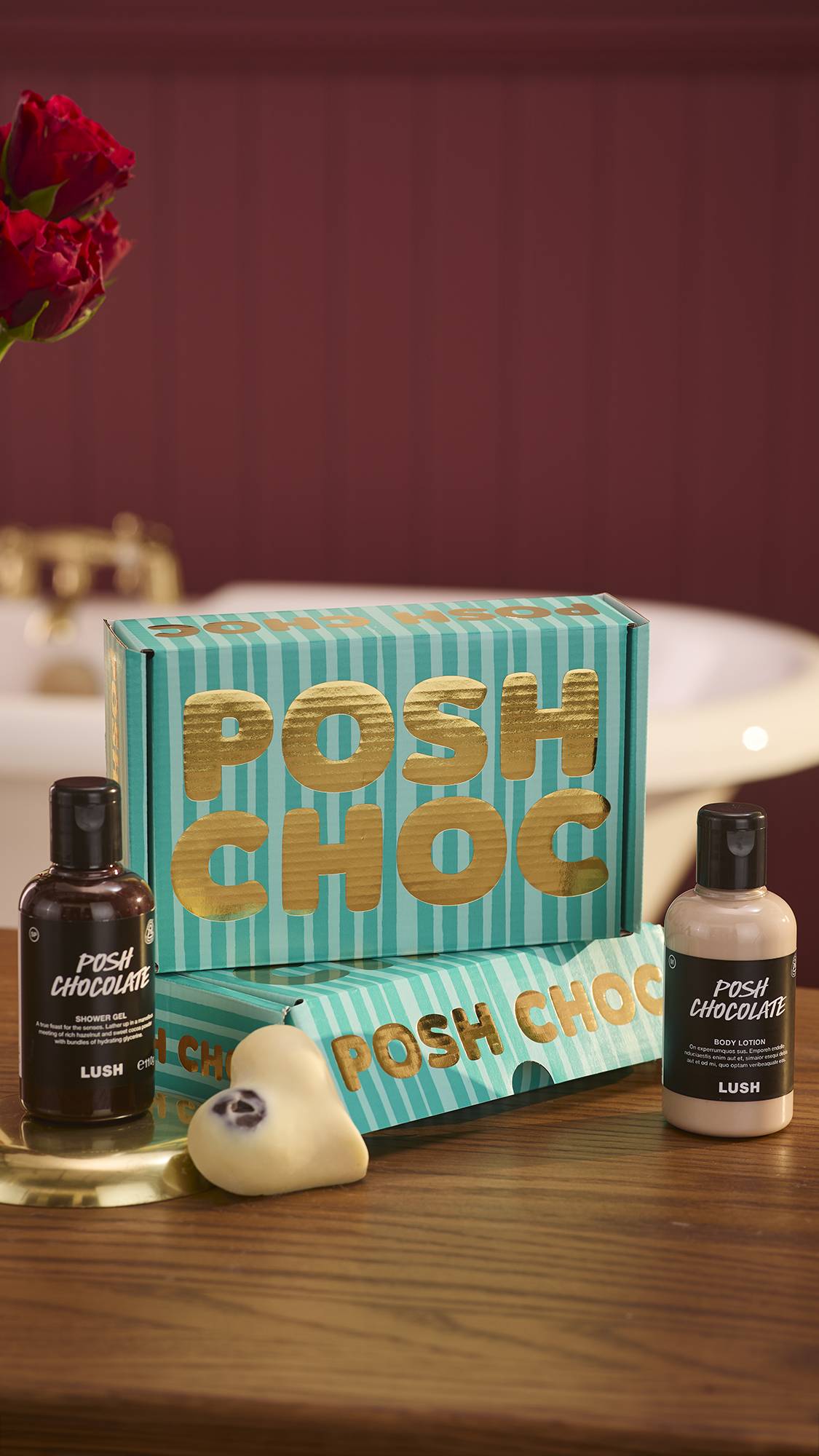 The image shows a dark, chocolate-coloured background with roses just visible on the left. Two stacked Posh Choc, teal-striped gift boxes sit on a wooden counter with the three, gourmand included products around them.