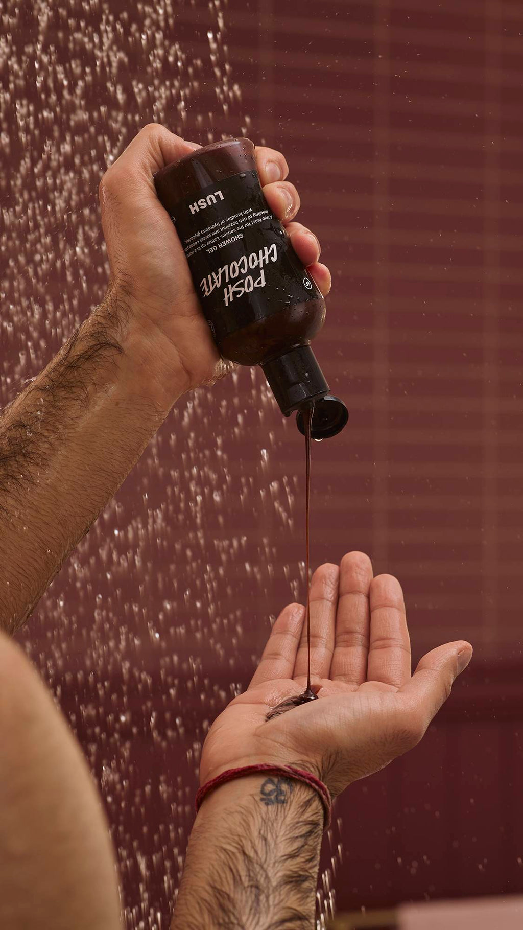 A close-up of the model's hands under running shower water as they squeeze the glossy Posh Chocolate shower gel from the product bottle into their palm. There is a chocolate-coloured background with running shower water.