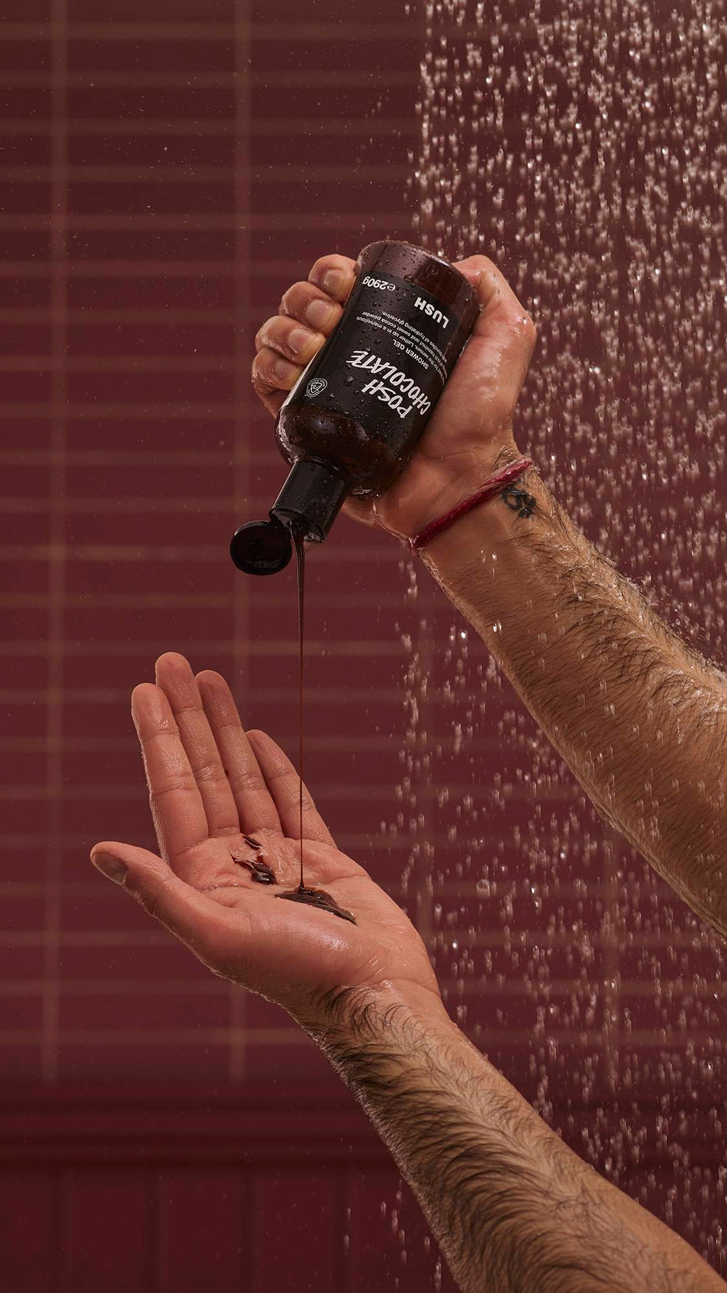 A close-up of the model's hands under running shower water as they squeeze the glossy, molasses-like Posh Chocolate shower gel from the product bottle into their hand. There is a chocolate-coloured background with running shower water.