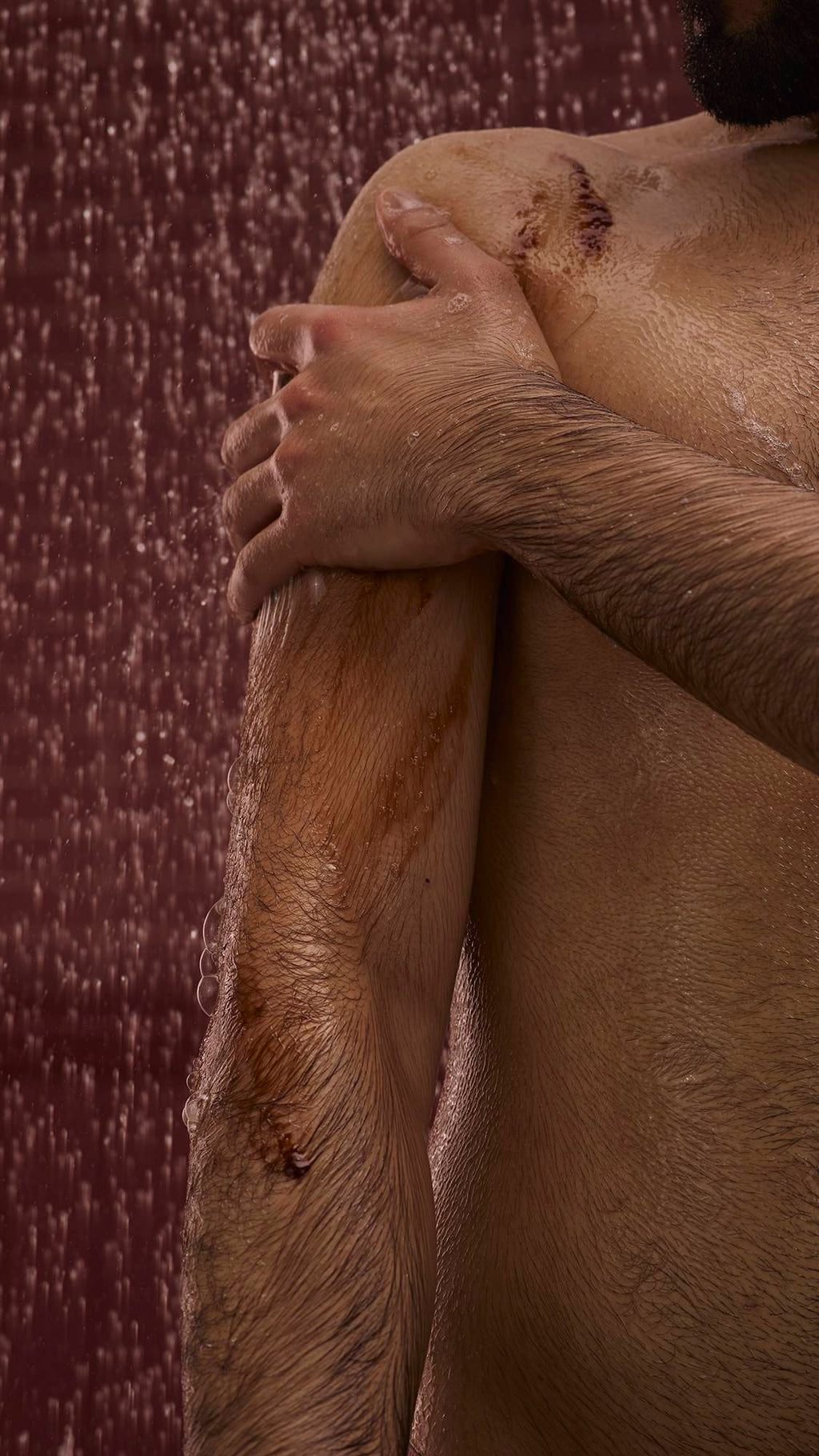 The image shows a dark, chocolate-coloured background with falling shower water. In the foreground, the model stands facing the camera as they lather up their shoulder with the Posh Chocolate shower gel to create a soft foam.