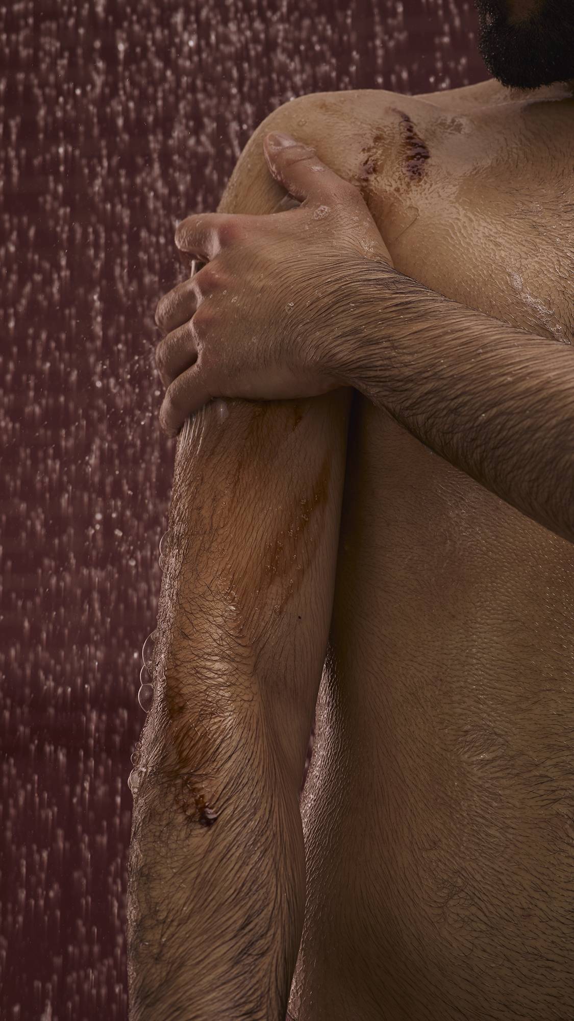 The image shows a dark, chocolate-coloured background with falling shower water. In the foreground, the model stands facing the camera as they lather up their shoulder with the Posh Chocolate shower gel to create a soft foam.