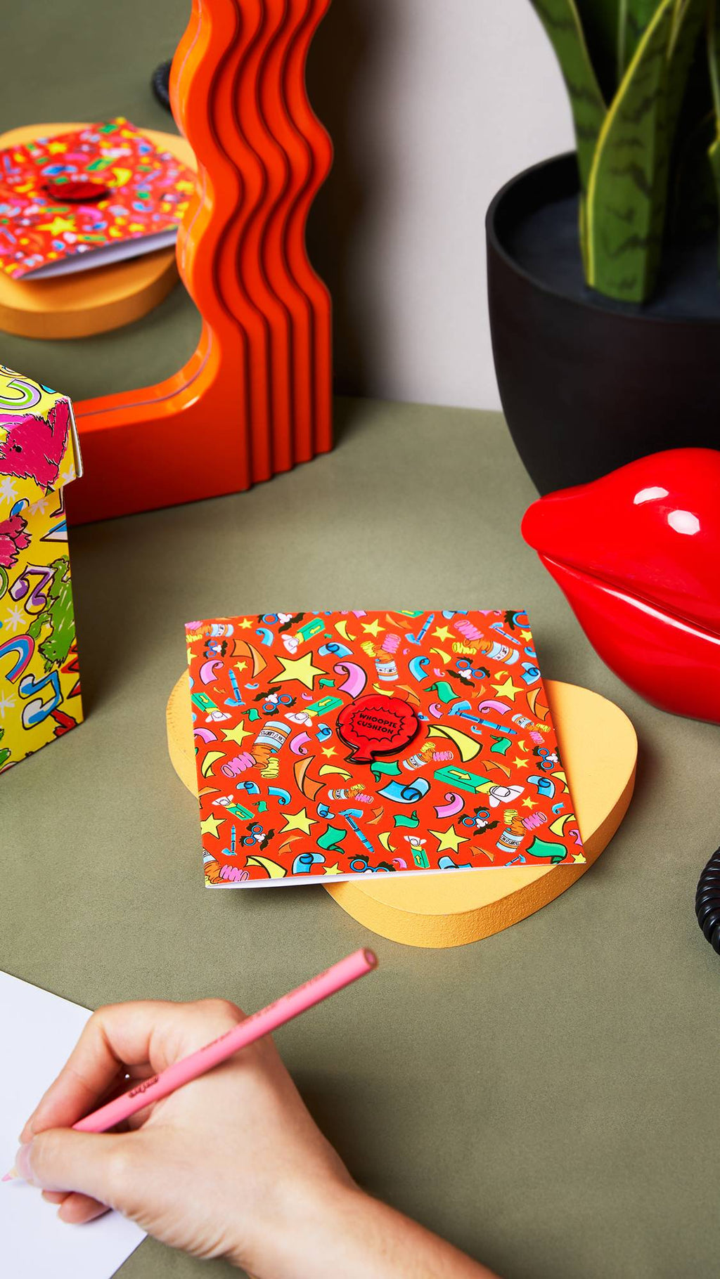 An angled, aerial image of the bright red Practical Joker greetings card sitting on a yellow wooden dish on a desk. There is a mirror, plant, and gift box sitting around the card as the model writes on the envelope with a pink pencil in the corner.