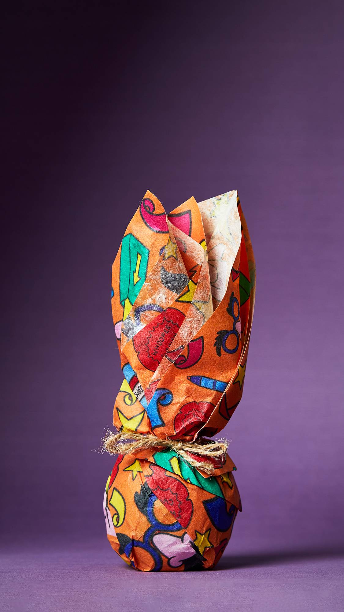 The image shows a deep, royal purple background. The foreground shows the bright orange, illustrated Practical Joker lokta paper wrapped neatly around a spherical gift, bunched and tied at the top with string.